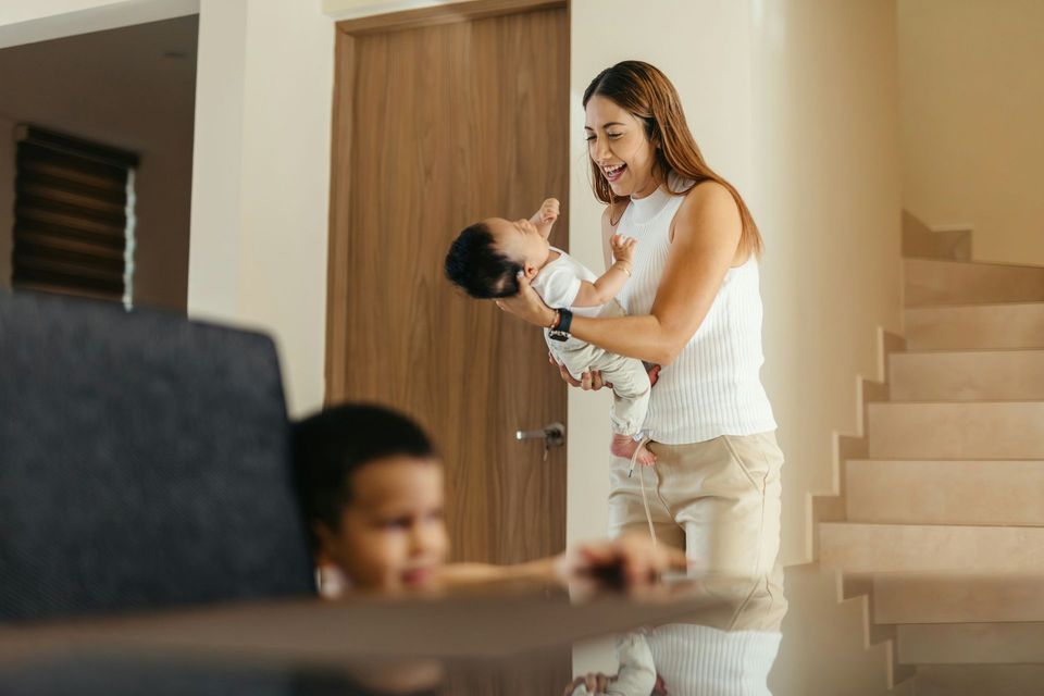 Woman holding baby, smiling, near staircase. Child watches from behind a table.