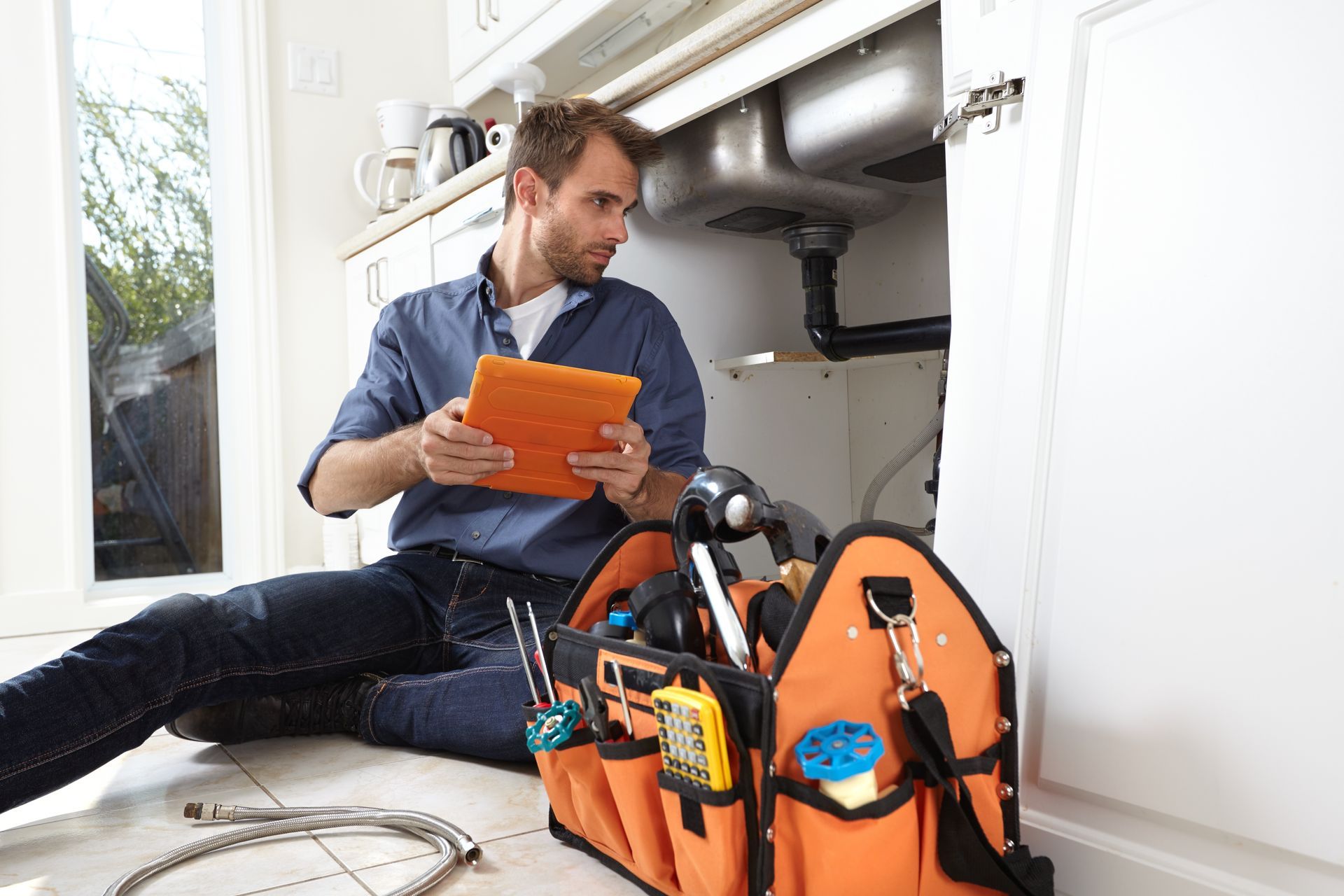 Plumber reading an orange manual beside a tool bag while working under a sink
