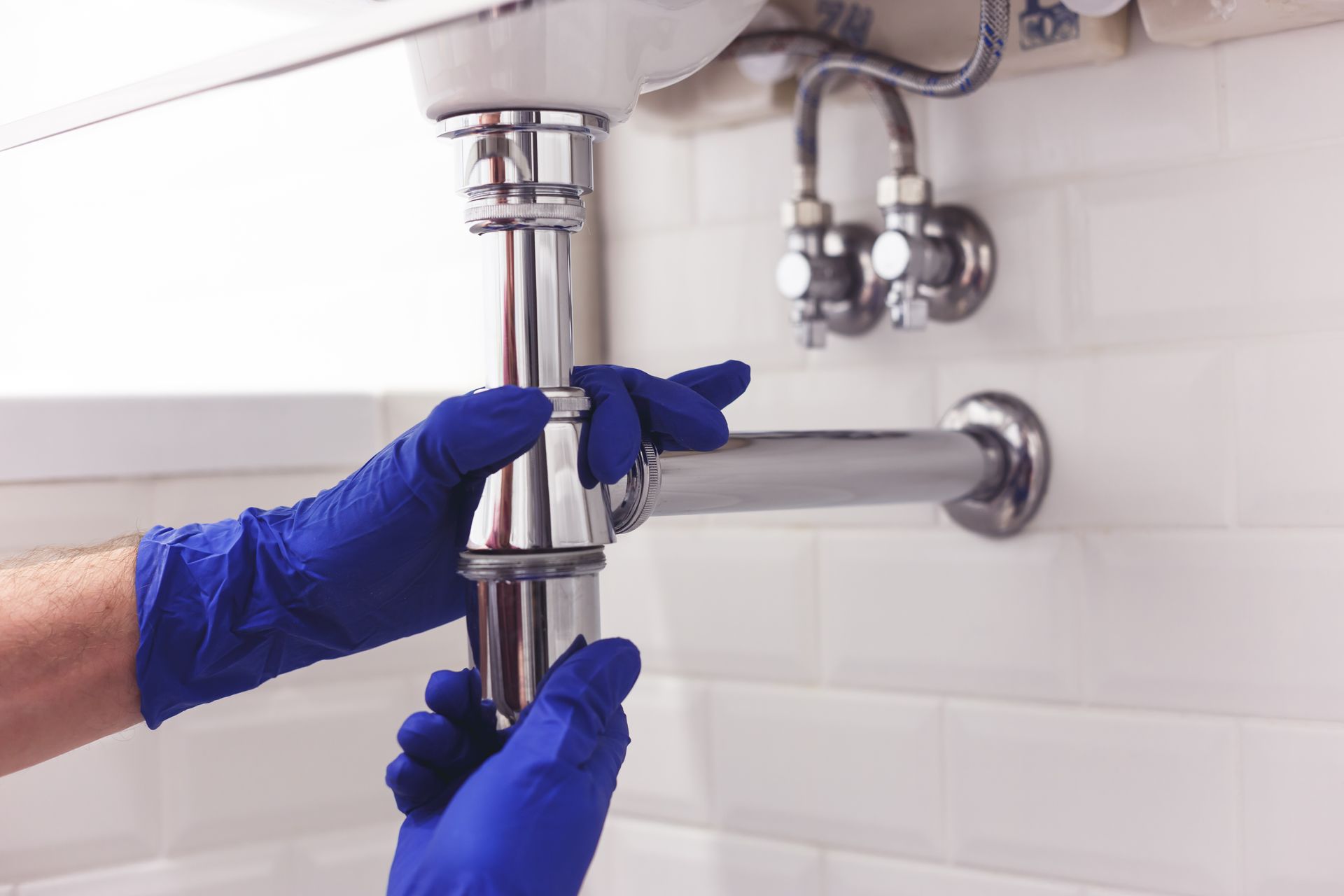 Blue-gloved hands tightening a bathroom sink drain pipe under a white basin