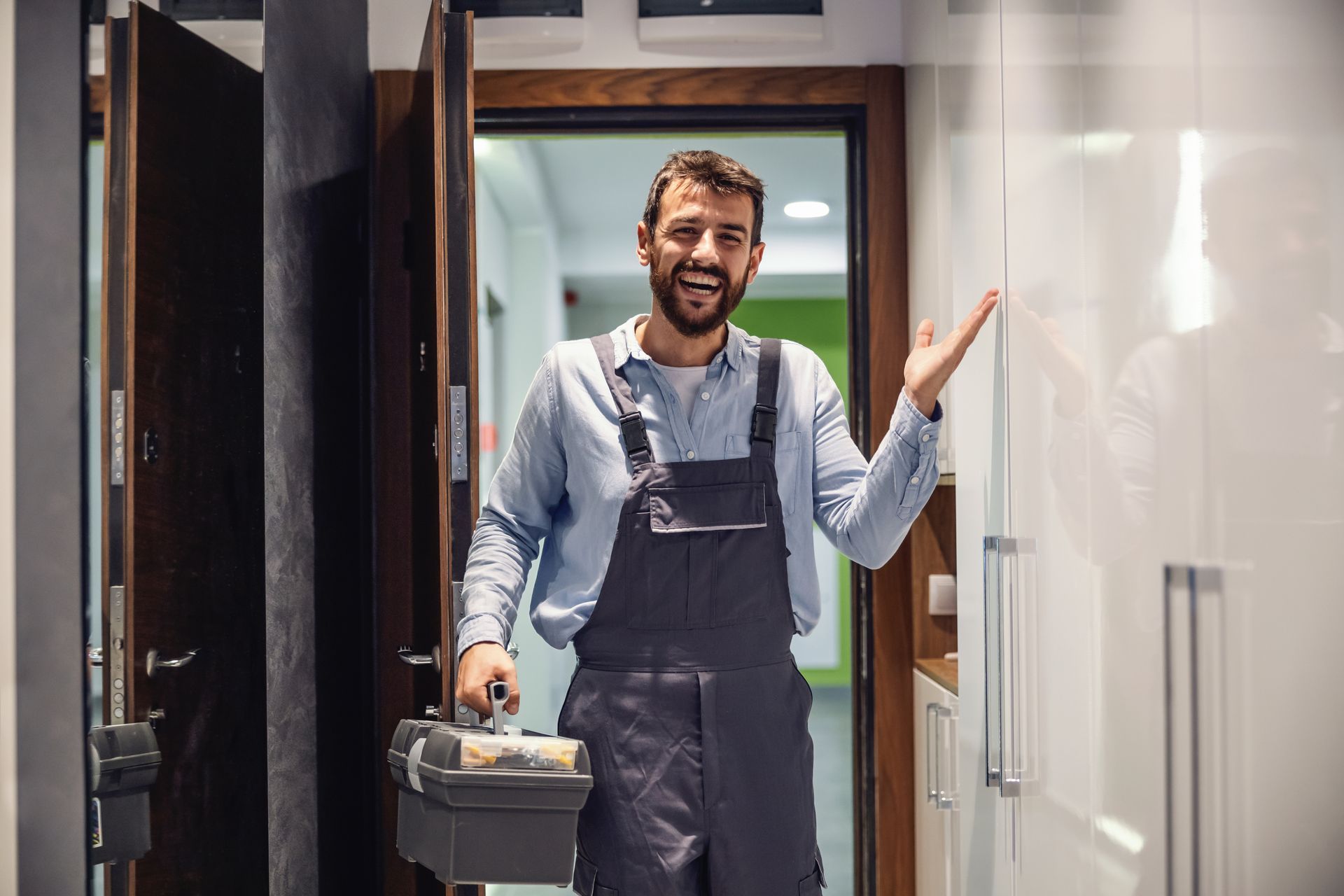 Smiling repair worker in blue overalls standing in a doorway, holding a toolbox and gesturing inside a modern home