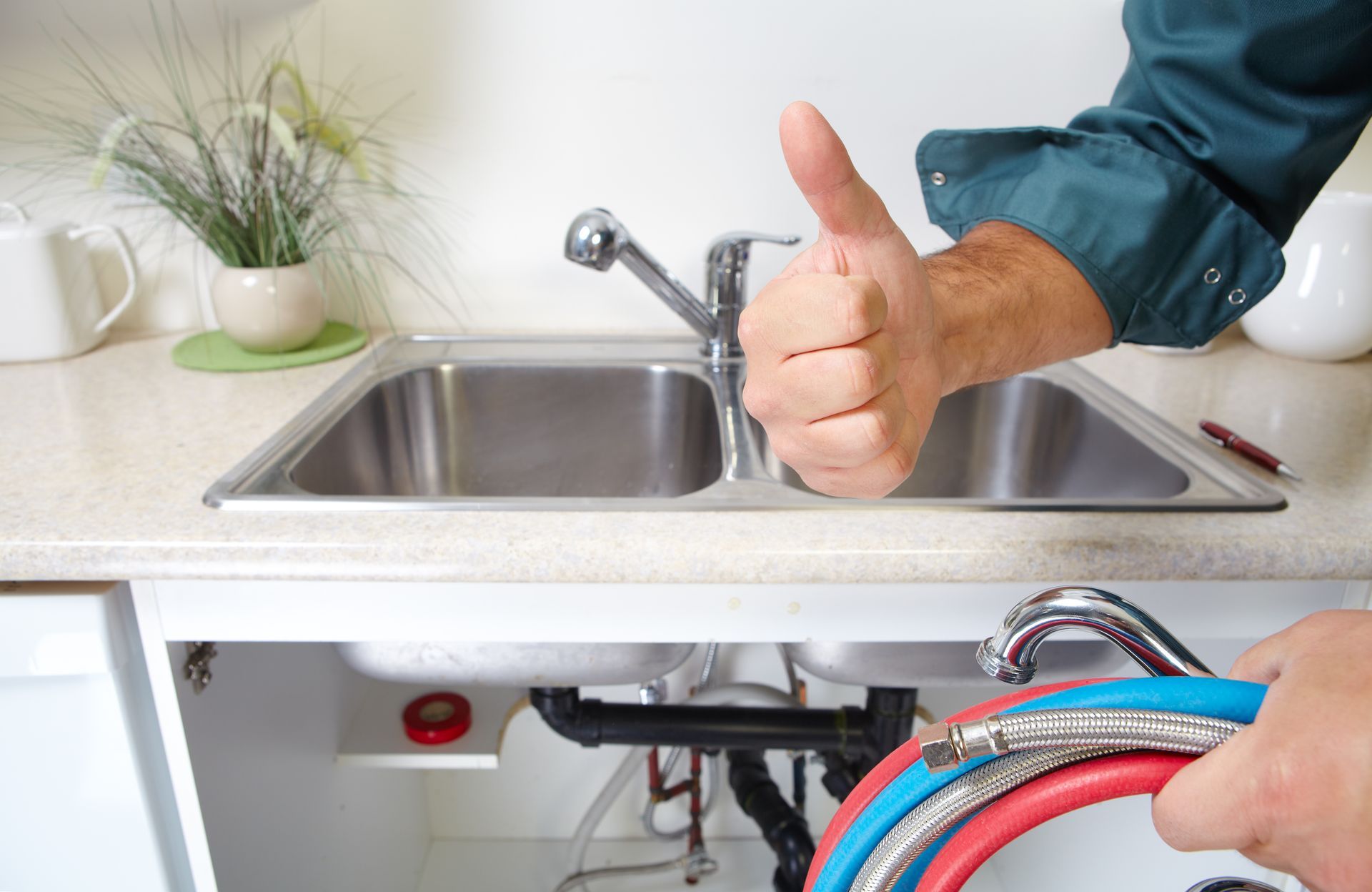 Hand giving thumbs-up over a kitchen sink while a plumber works under the counter with hoses exposed