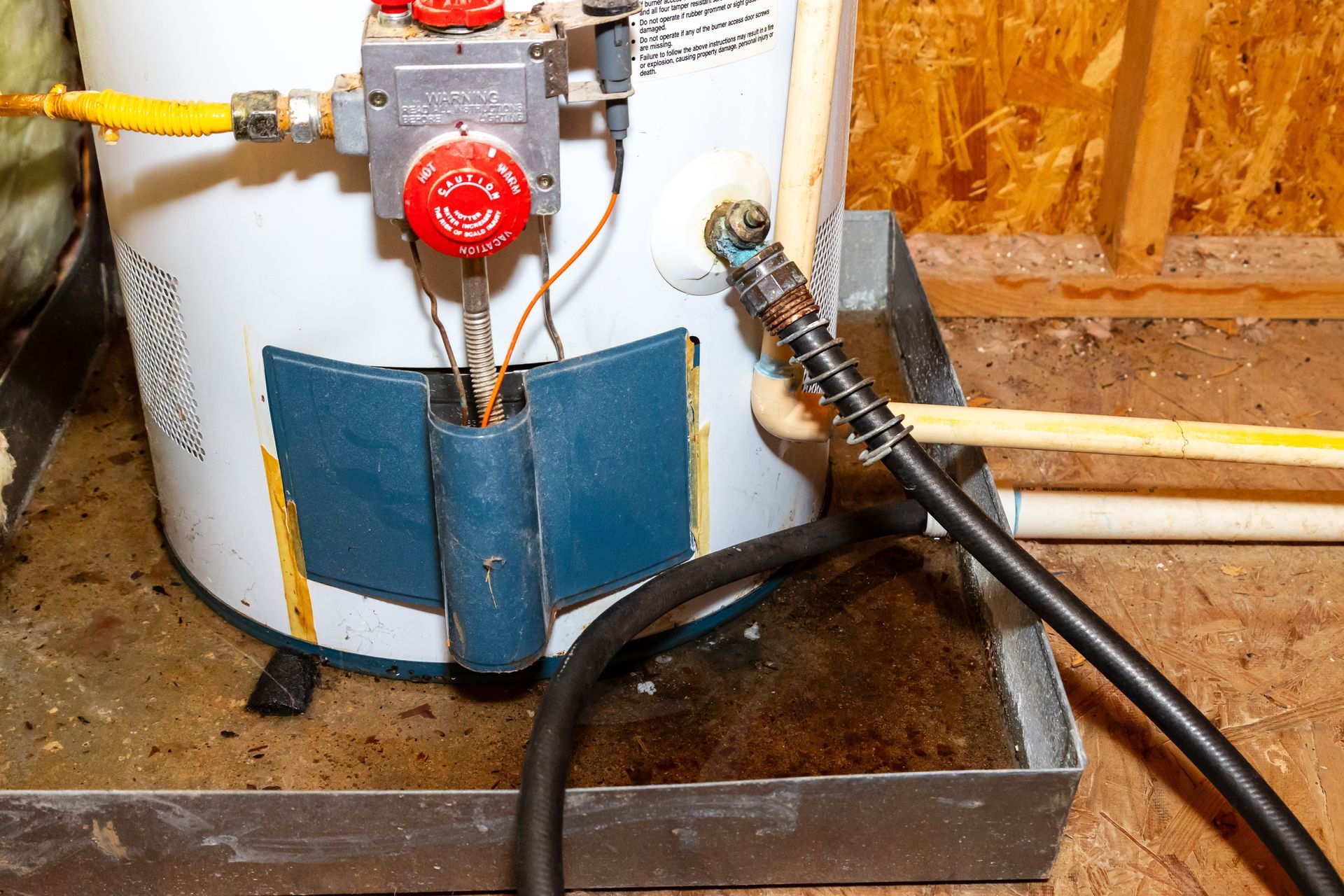 Water heater with a red valve and attached hoses, sitting in a shallow metal pan on a wooden floor.