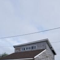 A gray, weathered shed with a brown shingled roof and a horizontal row of four windows under a cloudy sky.