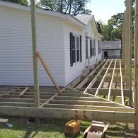 An L-shaped wooden deck frame under construction alongside a white single-story house with black-shuttered windows.