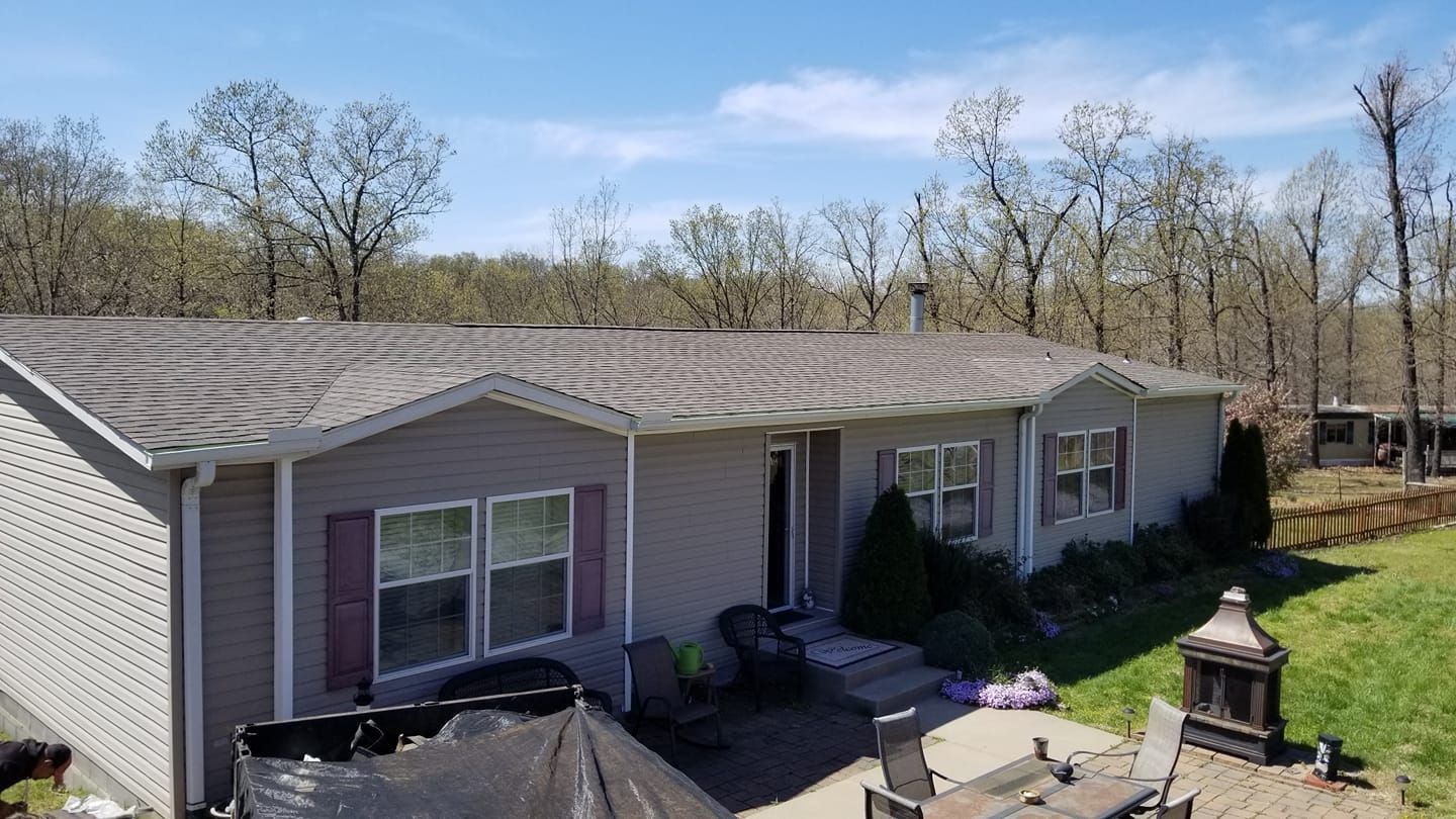 A gray, single-story ranch-style home sits in a sunny yard with a patio and a wooded backdrop.