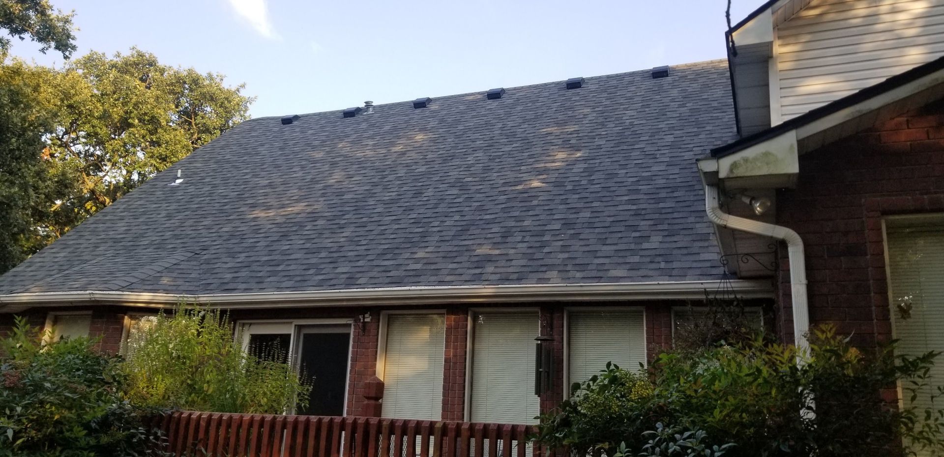 A house with a brick exterior, white trim, and a shingled roof under a clear blue sky, partially obscured by green trees.