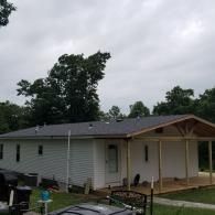 A single-story white house with light grey siding, a dark shingled roof, and an unfinished wooden porch under cloudy skies.
