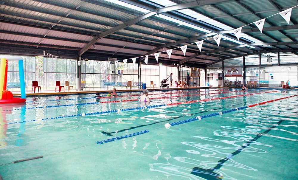 Indoor swimming pool with lane markers, red chairs, and colorful pool noodles under a metal roof