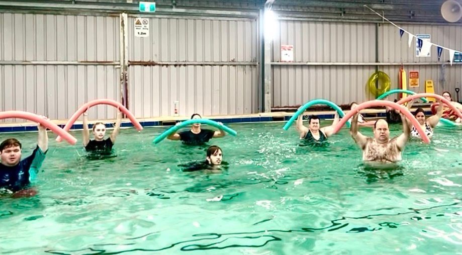 People doing aqua aerobics in a pool, including a man holding red foam dumbbells in the foreground