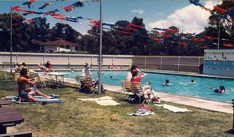 Outdoor pool with swimmers, people lounging on grass, and red umbrellas under a sunny sky