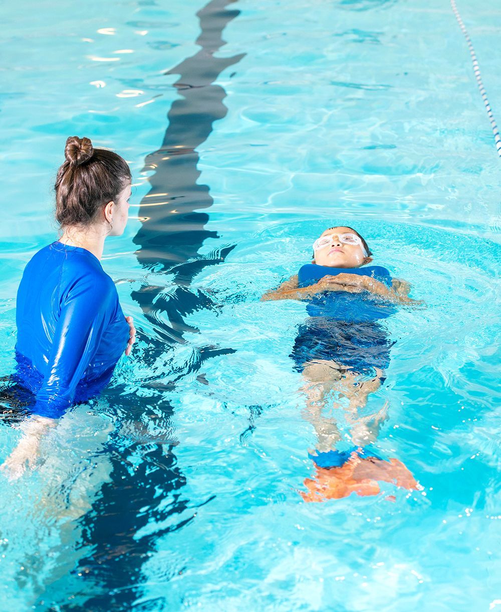 Two people in a swimming pool, one instructing as the other floats on their back.