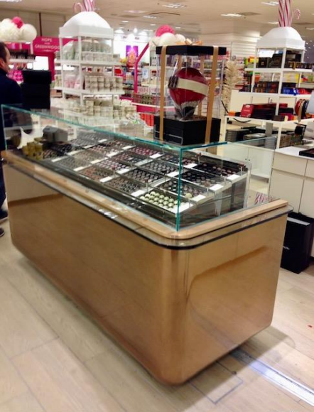 Vitrine de chocolats dans un magasin très éclairé, avec un dessus en verre et des côtés en bois courbés.