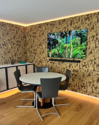 Coin repas avec table ronde, quatre chaises, TV, papier peint à motifs dorés et parquet.