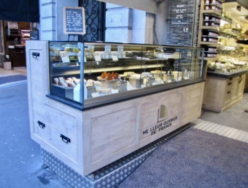 Vitrine à fromages devant un magasin, remplie de fromages variés. Extérieur en bois clair, plateau en verre.