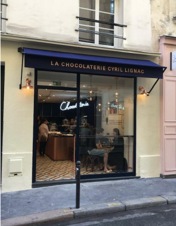 Une chocolaterie parisienne avec un auvent bleu, des vitrines en verre et une enseigne néon « Chocolat ».