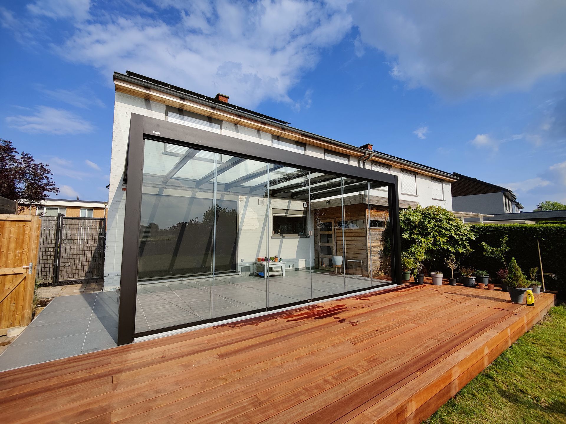 Une véranda aux parois de verre avec une terrasse en bois, attenante à une maison, sous un ciel bleu.