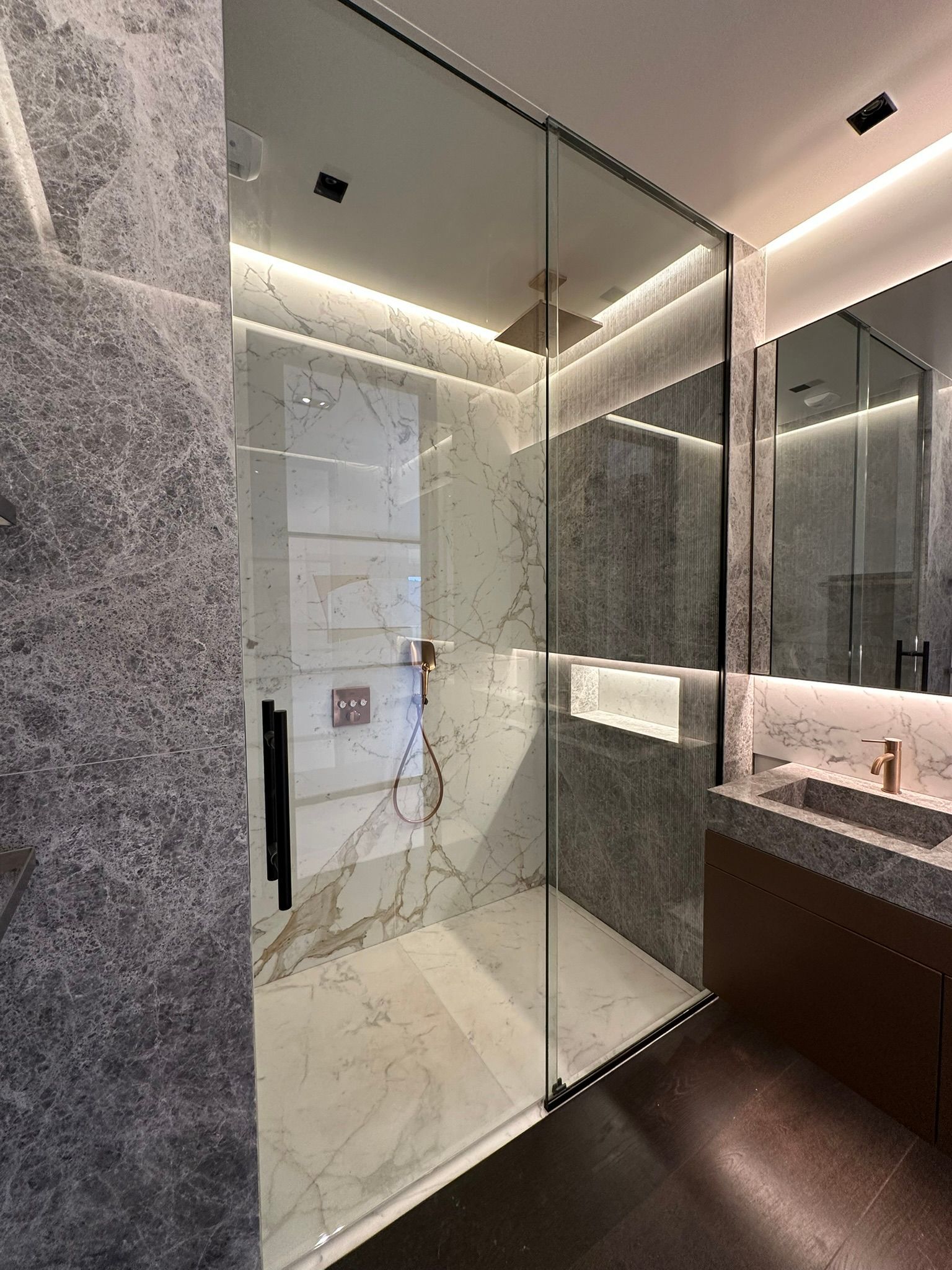 Salle de bain moderne avec douche en verre, murs en marbre, vanité en bois foncé et éclairage d'ambiance.