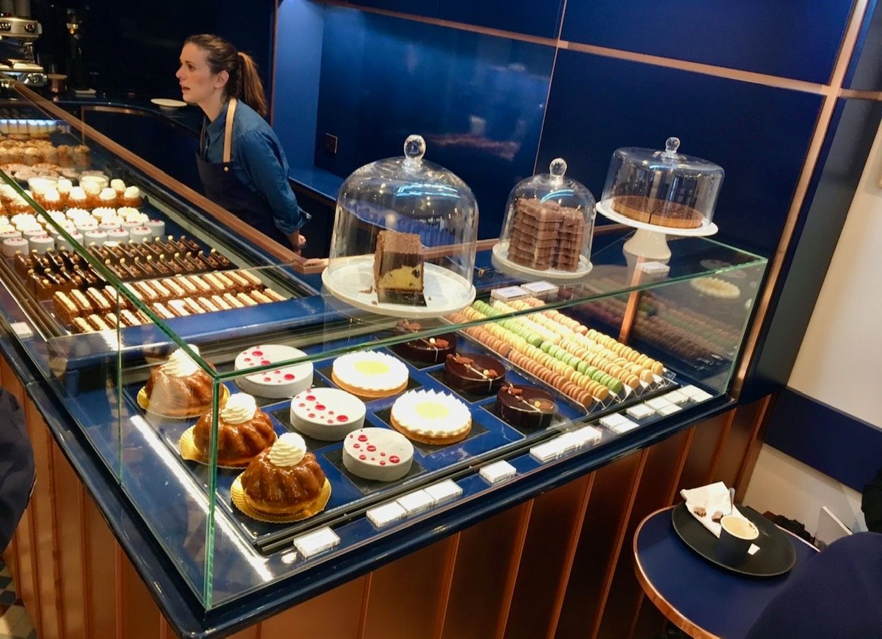 Présentoir de pâtisseries dans une boutique avec gâteaux, tartes et un serveur derrière le comptoir.