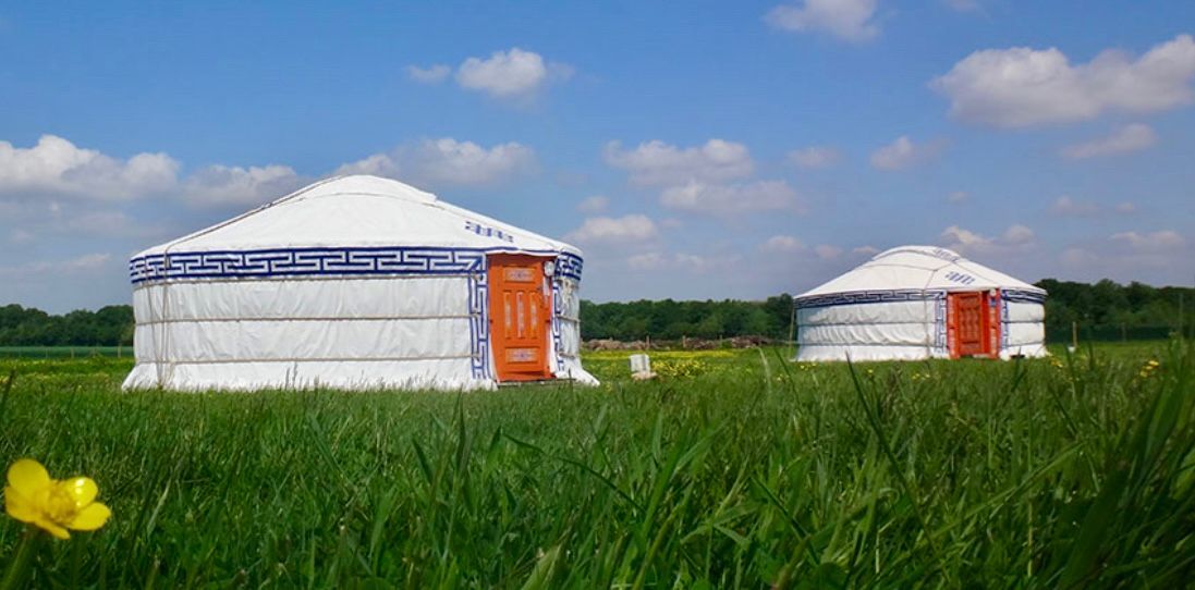 Deux yourtes blanches avec des portes orange dans un champ vert sous un ciel bleu avec des nuages.