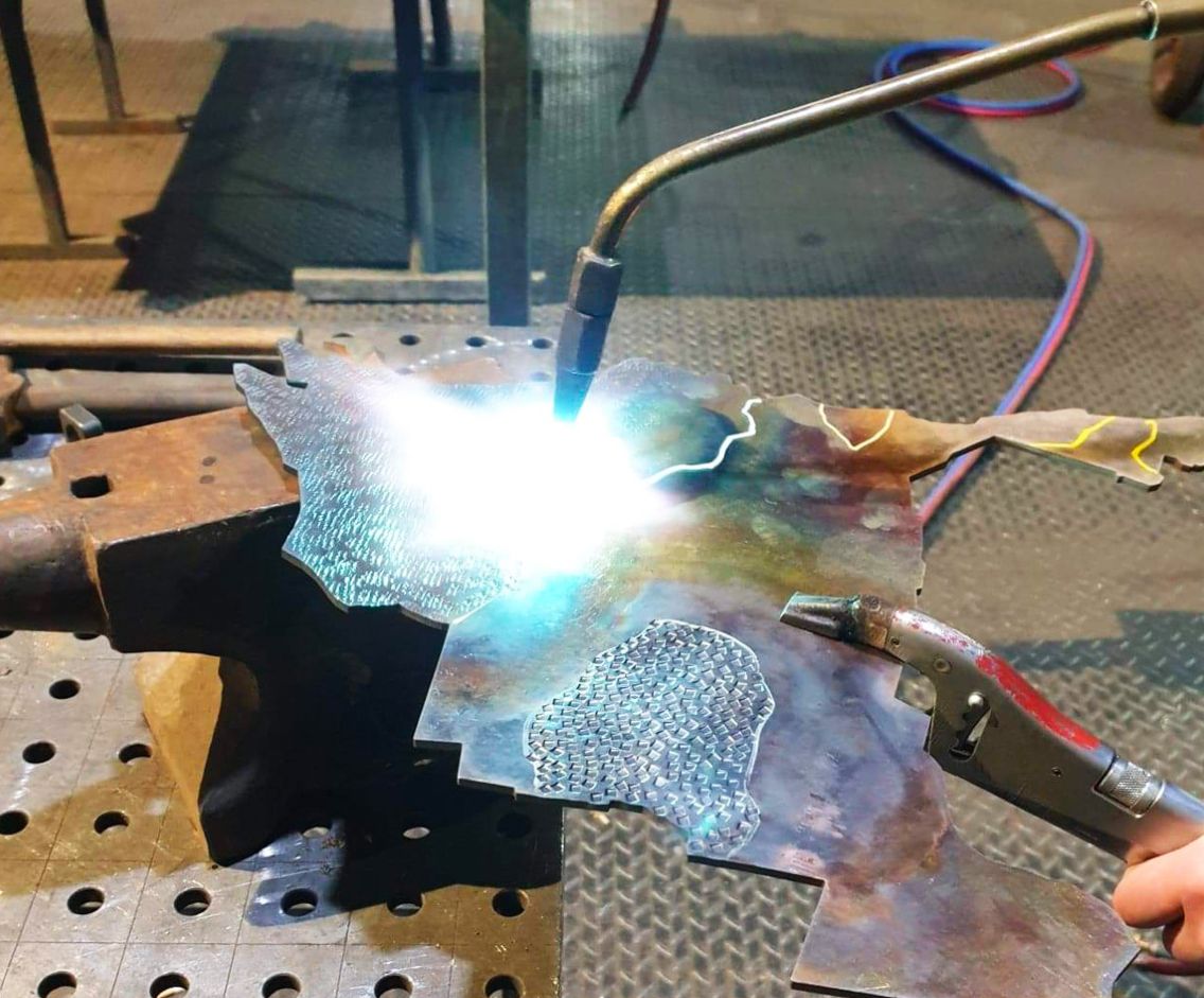 Une personne soude une pièce de métal avec un chalumeau, créant une étincelle bleue et blanche brillante sur un établi.