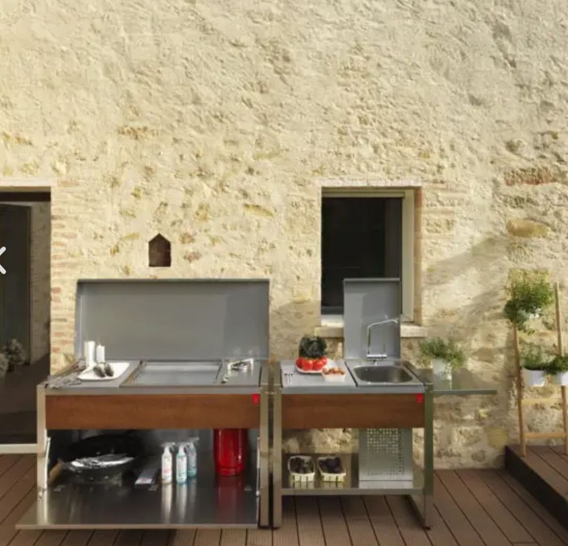 Cuisine extérieure avec grill, évier et rangement sur une terrasse en bois, contre un mur en pierre.