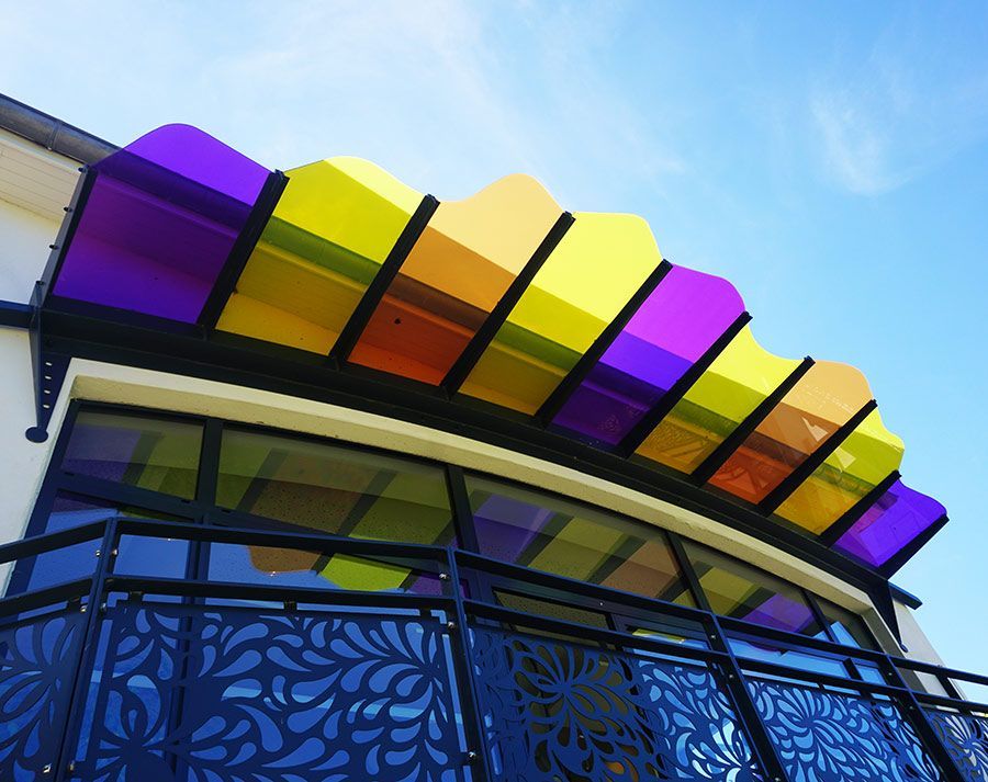 Auvent coloré au-dessus de l'entrée d'un bâtiment avec des panneaux violets, jaunes et orange, ciel bleu.