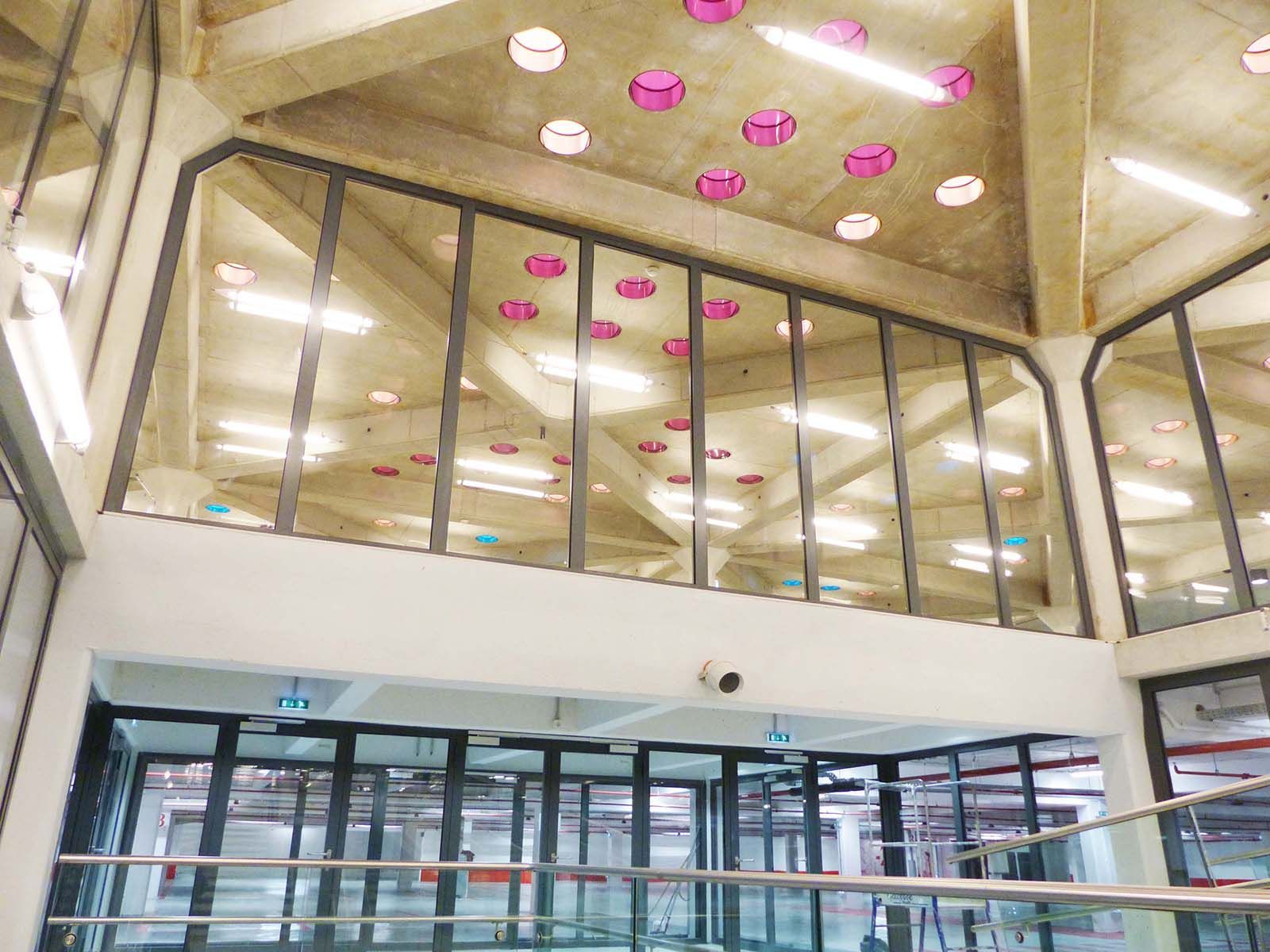 Vue intérieure d'un bâtiment moderne avec des murs en verre, un plafond en béton et des lumières roses décoratives.