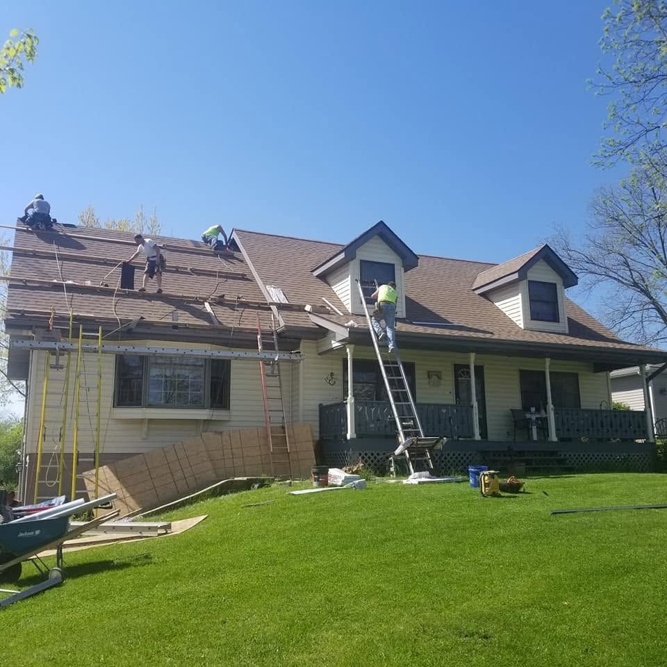 Hand using a nail gun to fasten roof shingles on a rooftop, with another worker blurred in the background