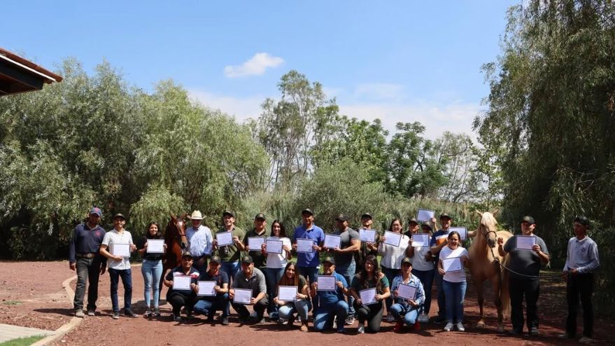 Grupo de personas posando al aire libre en un jardín; algunas sostienen certificados y, detrás, se ven árboles y un cielo azul.