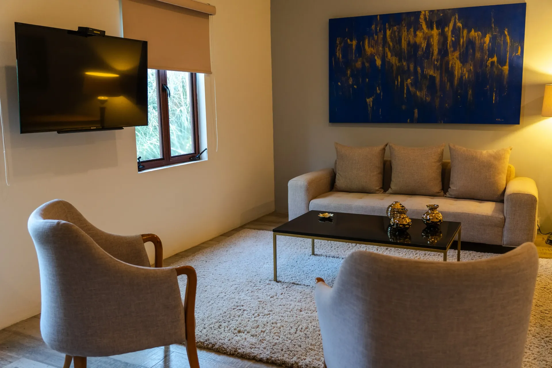 Sala de estar moderna con televisor montado en la pared, sofá beige, dos sillones y un cuadro abstracto azul