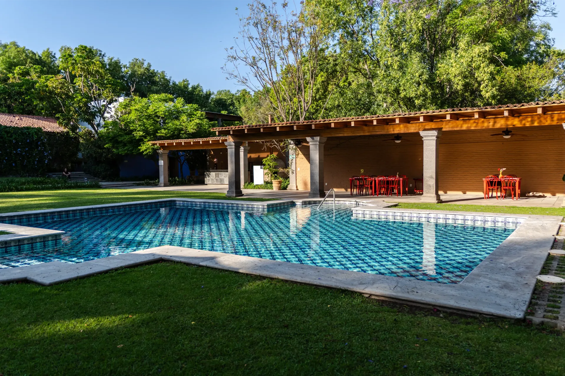 Piscina al aire libre junto a un patio cubierto con sillas rojas y césped verde bajo los árboles