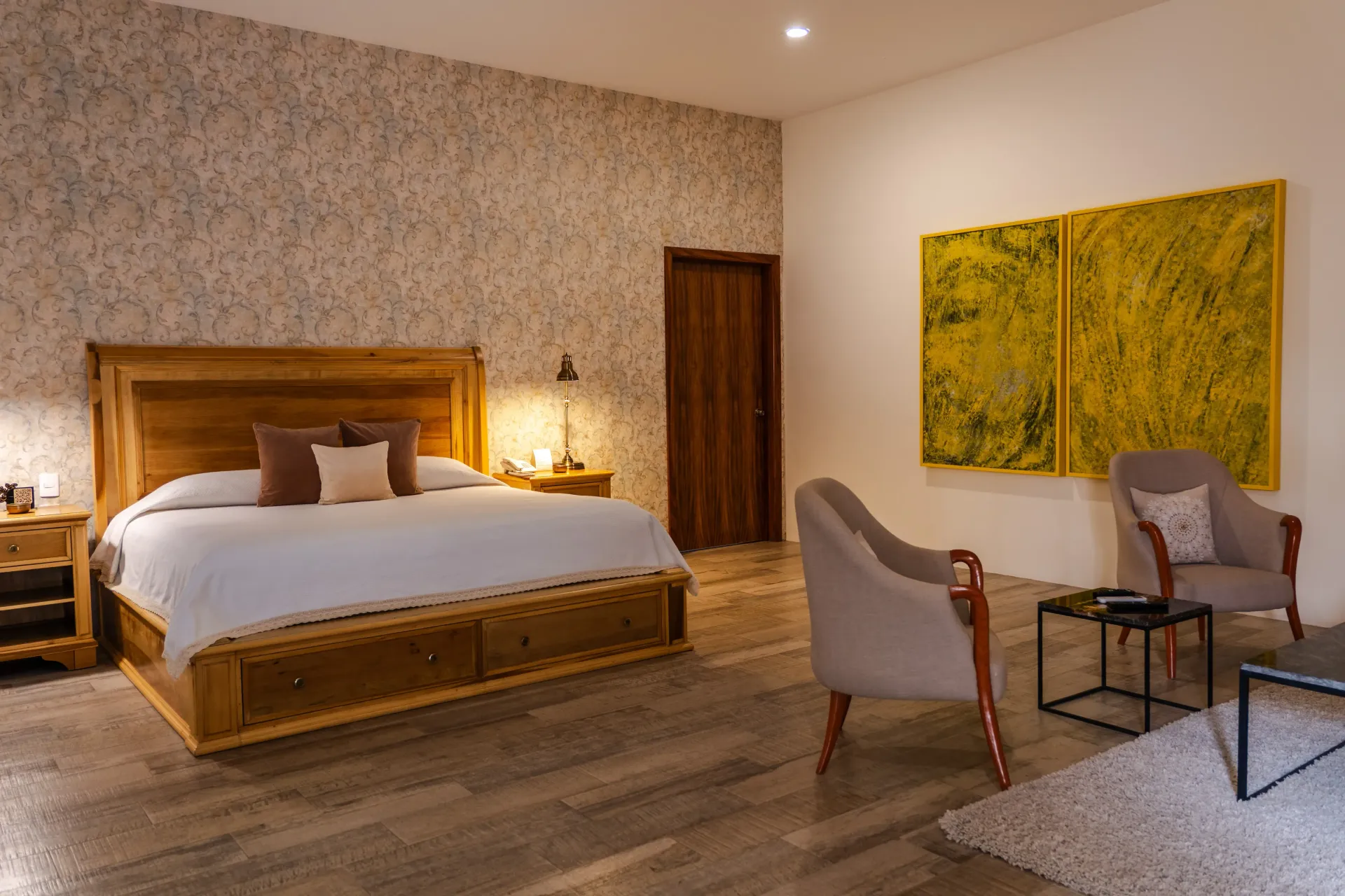 Dormitorio amplio con cama de madera, papel pintado estampado, cuadros amarillos y zona de estar