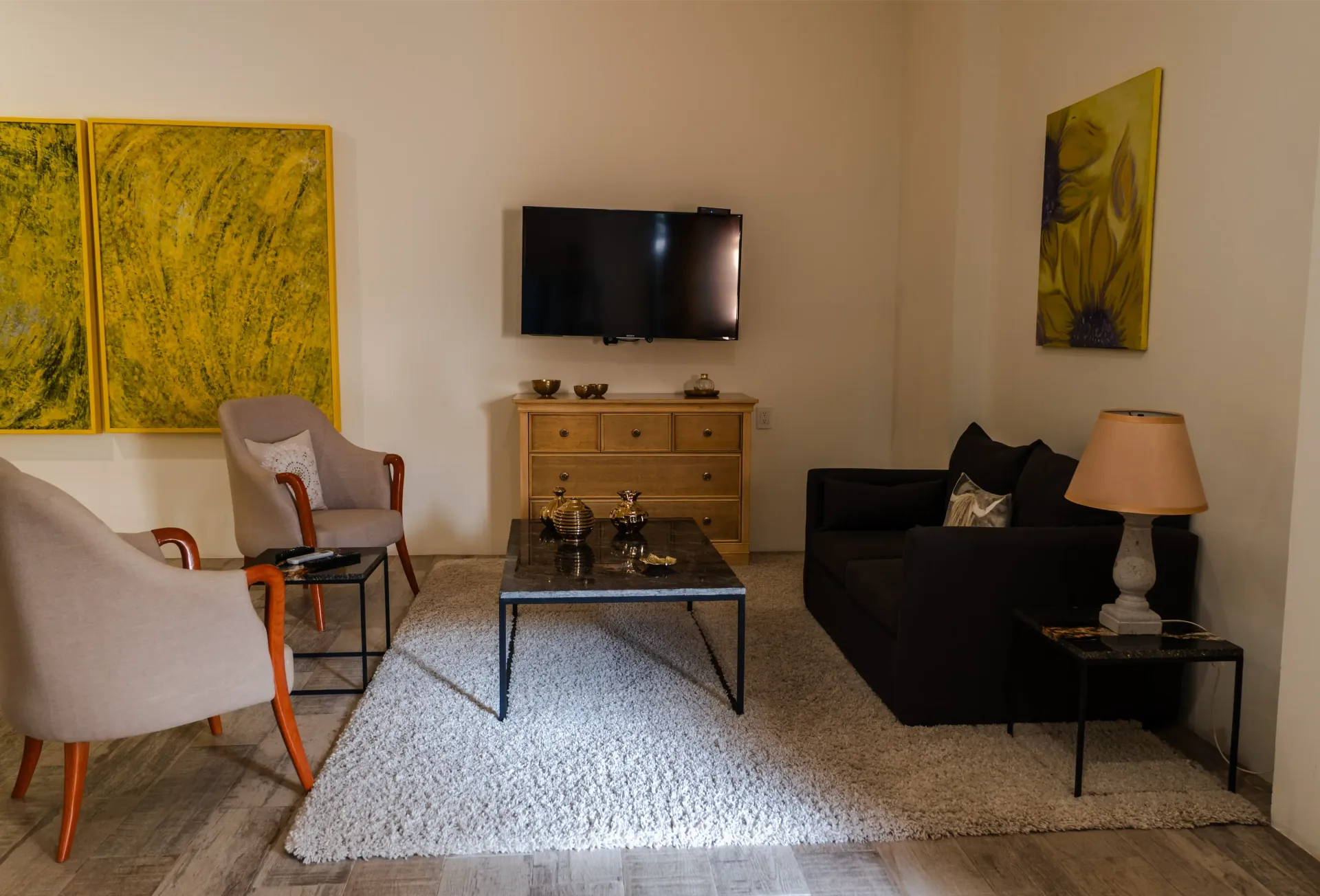 Salón moderno con televisor, sofá negro, sillones blancos, mesa de centro y cuadros amarillos