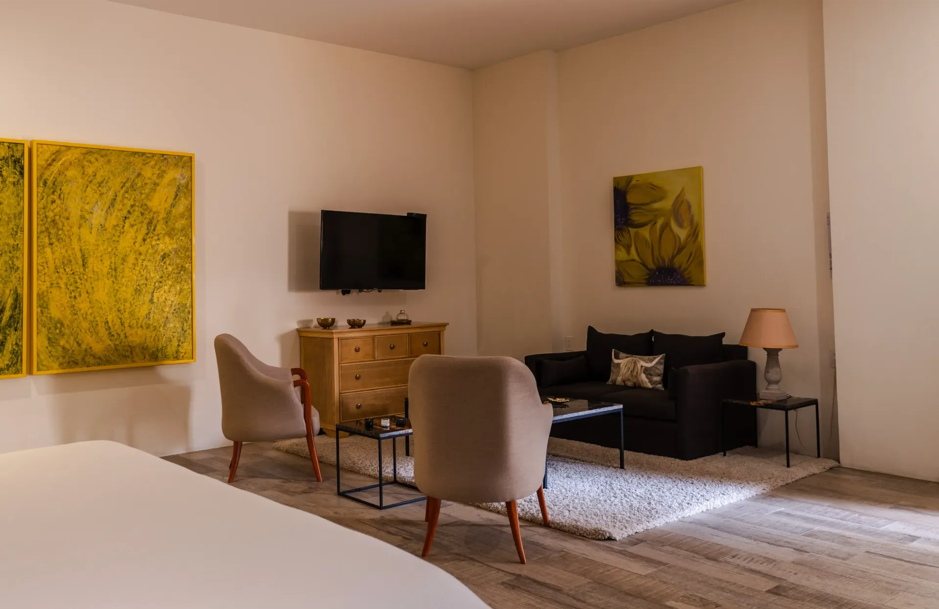 Sala de estar moderna con sillones beige, sofá negro, televisor montado en la pared, obra de arte amarilla y una lámpara.