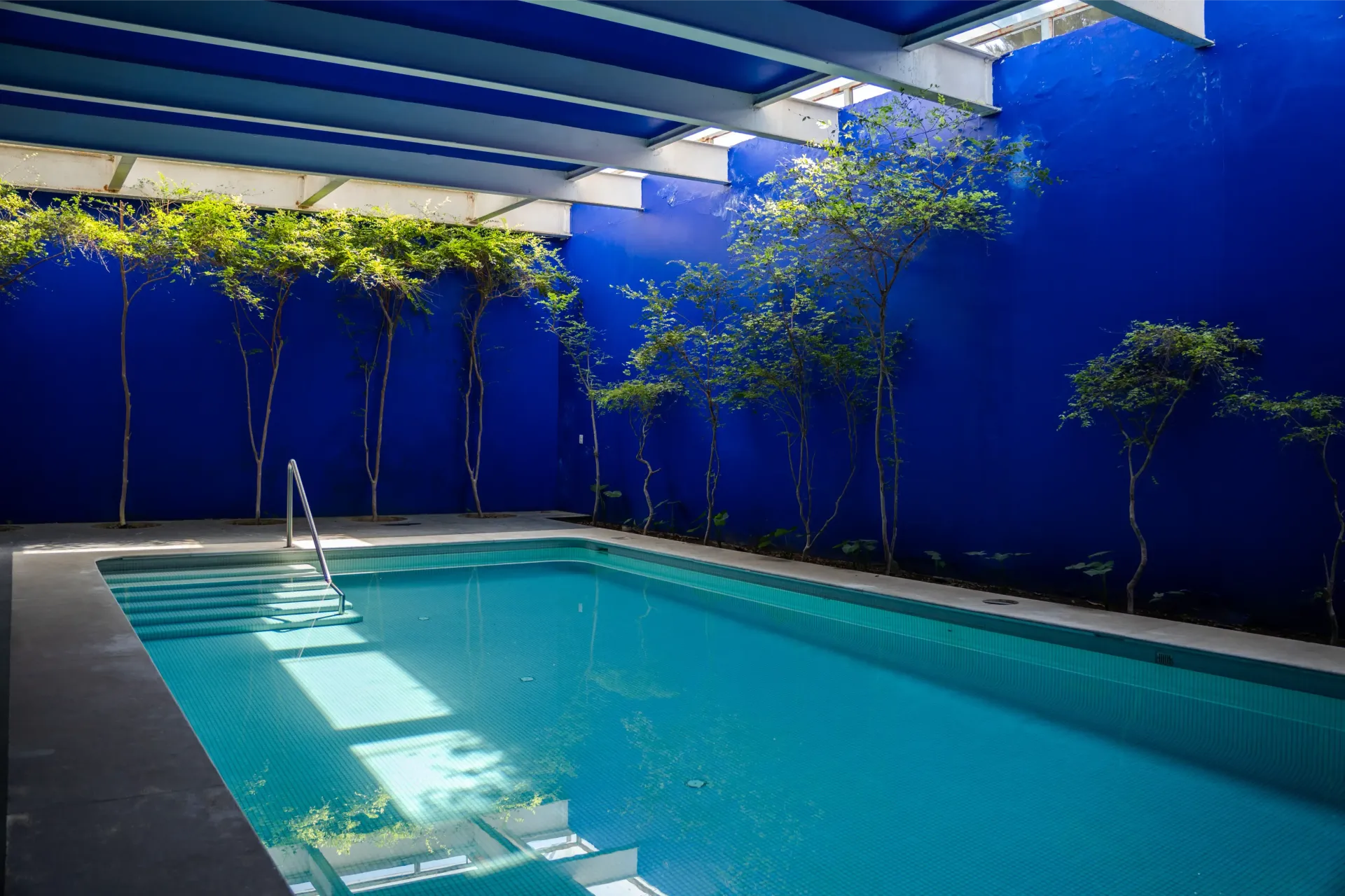 Piscina cubierta al aire libre con paredes azules, vigas blancas y plantas en macetas a lo largo de la tarima
