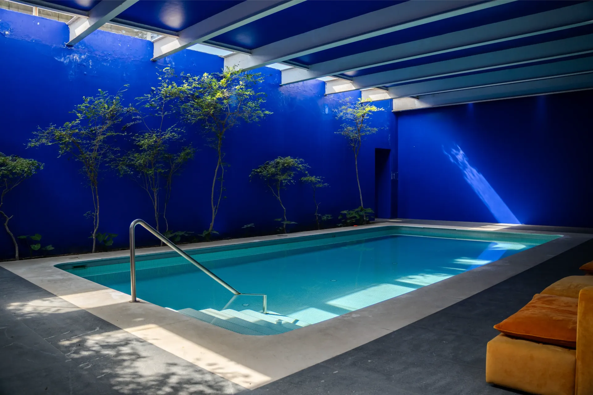 Piscina cubierta moderna con paredes azules, vigas bajo un tragaluz y tumbonas naranjas