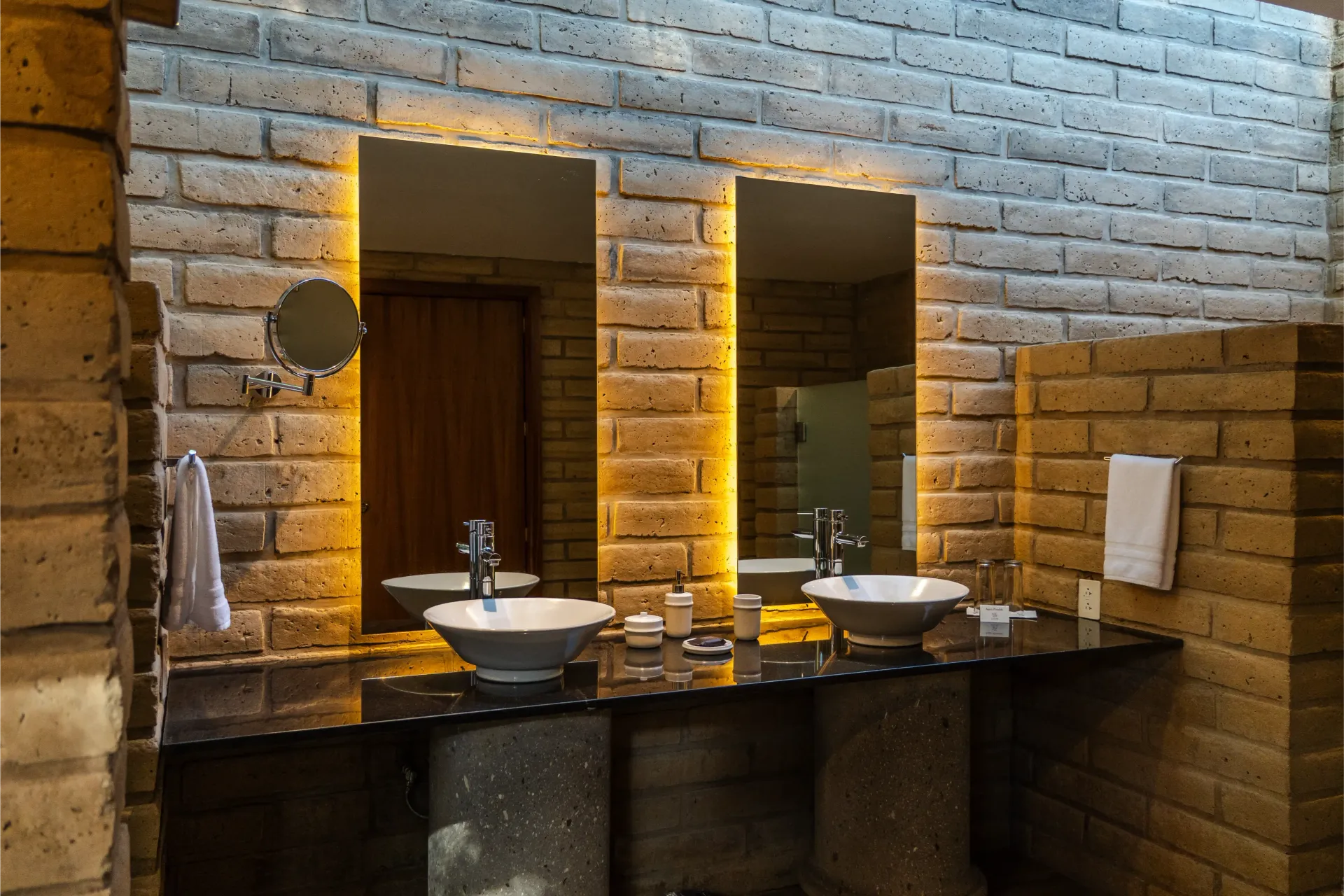 Baño con paredes de ladrillo, dos lavabos, espejos e iluminación cálida retroiluminada