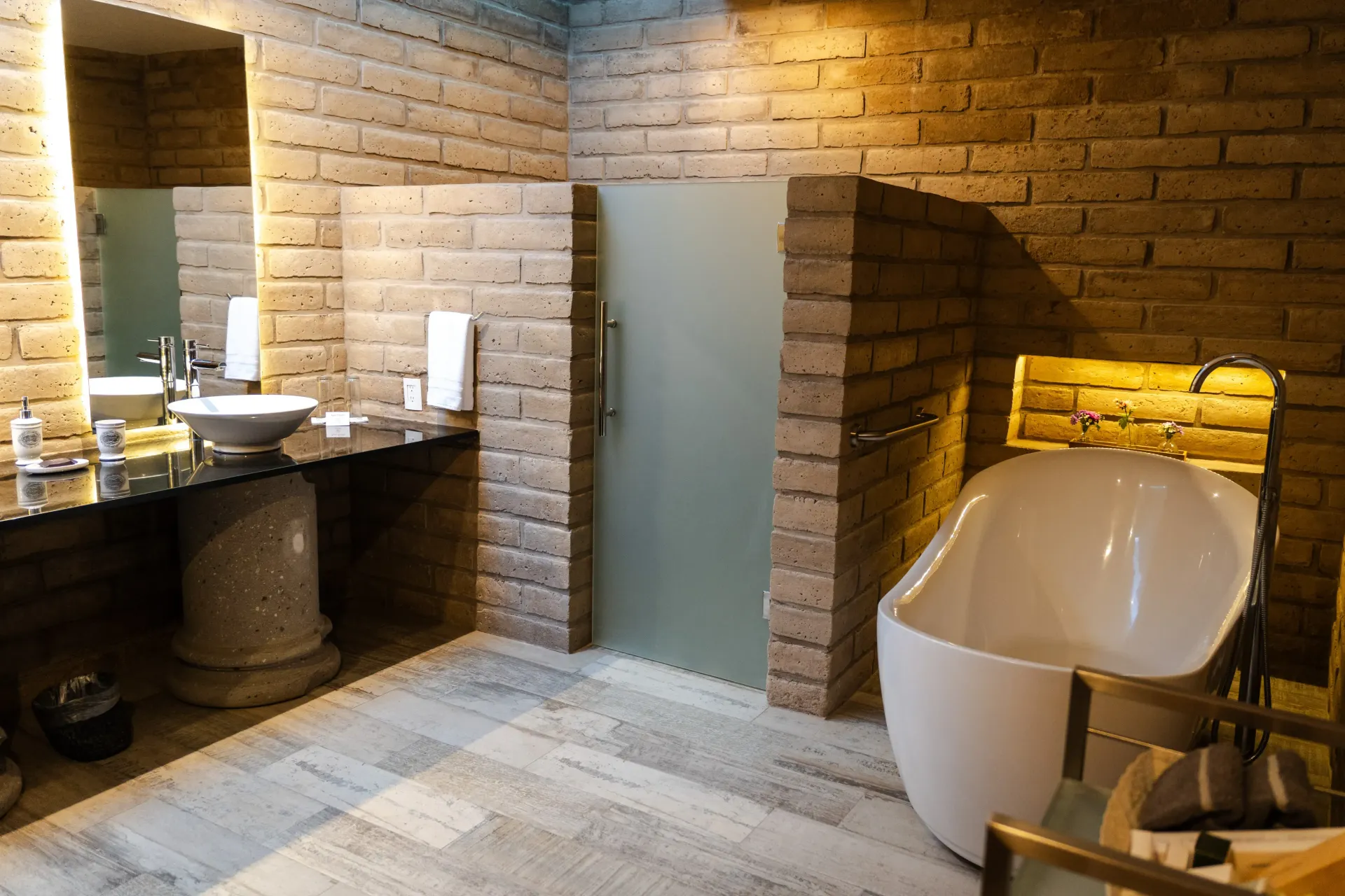 Baño moderno con paredes de piedra, tocador con lavabo doble, ducha con mampara esmerilada y bañera exenta blanca