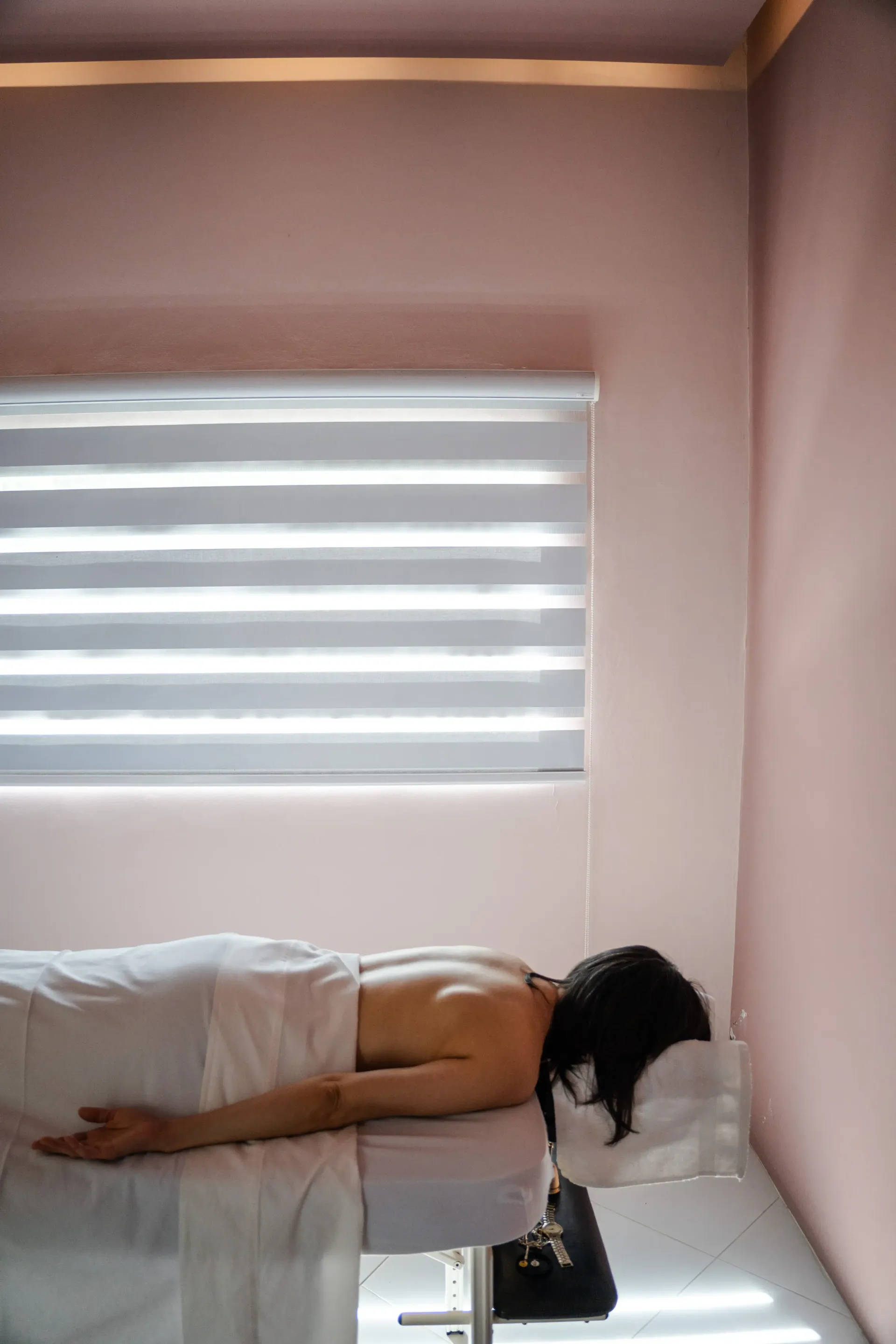 Persona tumbada boca abajo en una cama, en una habitación rosa junto a una ventana con persianas.