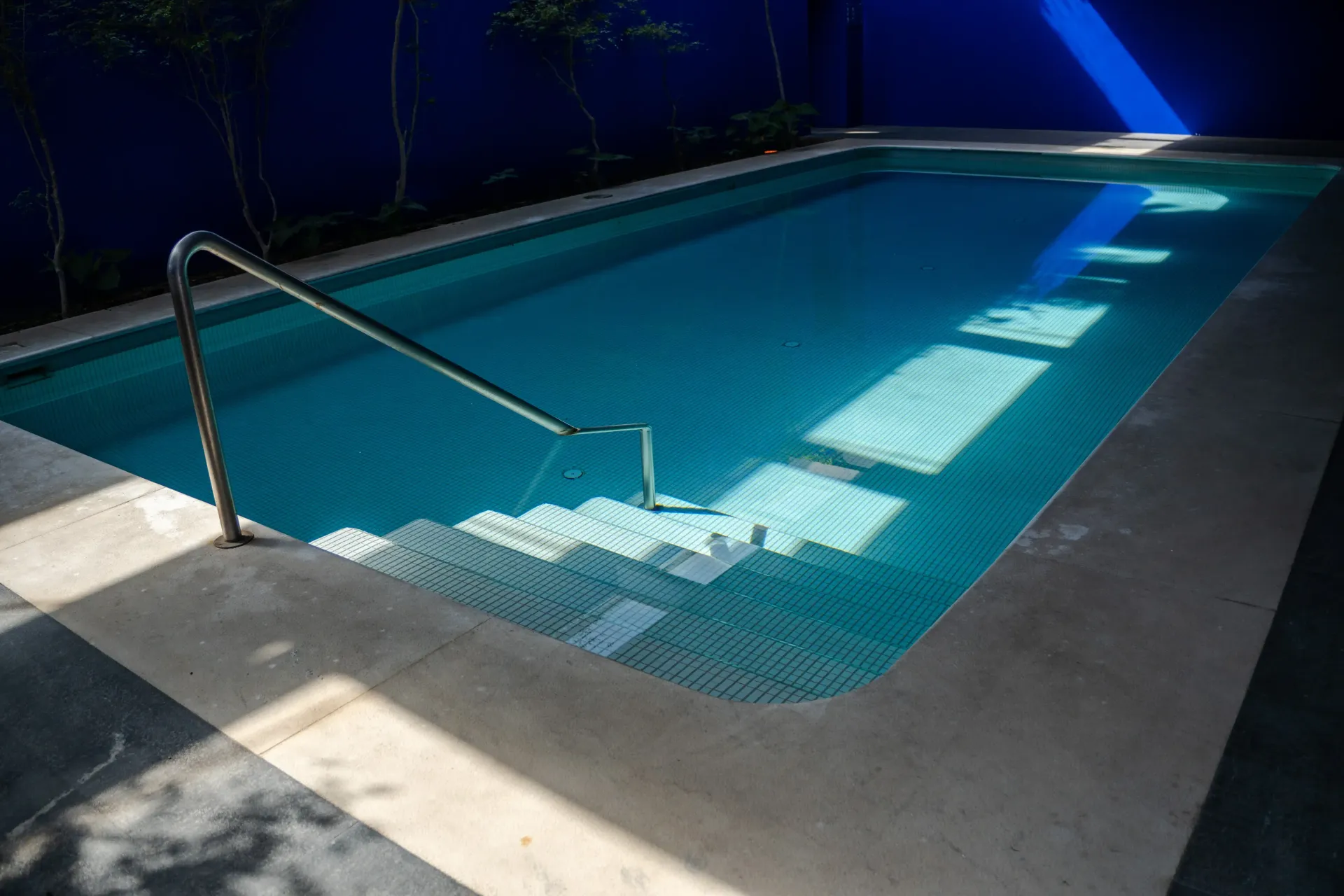 Piscina vacía con agua azul, una barandilla metálica y la luz intensa del sol proyectando sombras sobre la terraza