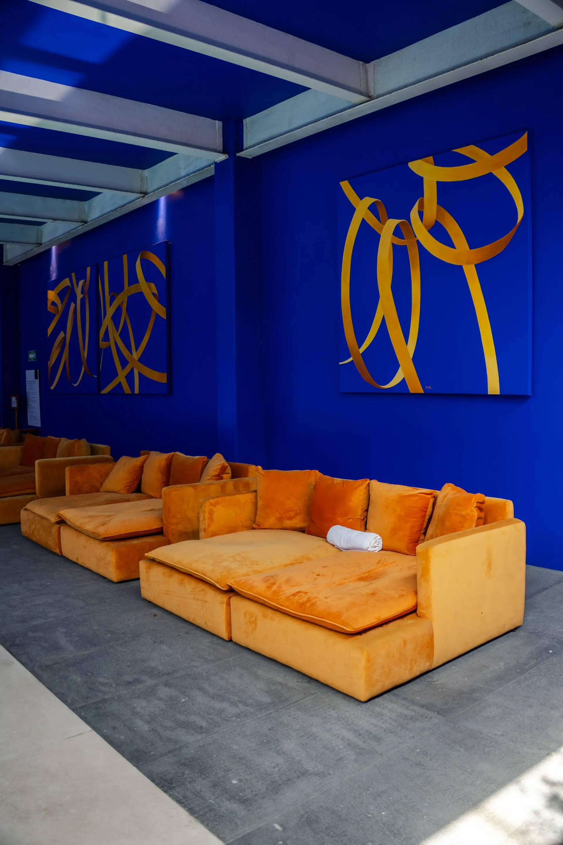 Sofás naranjas junto a una pared azul, con arte abstracto dorado bajo un toldo azul