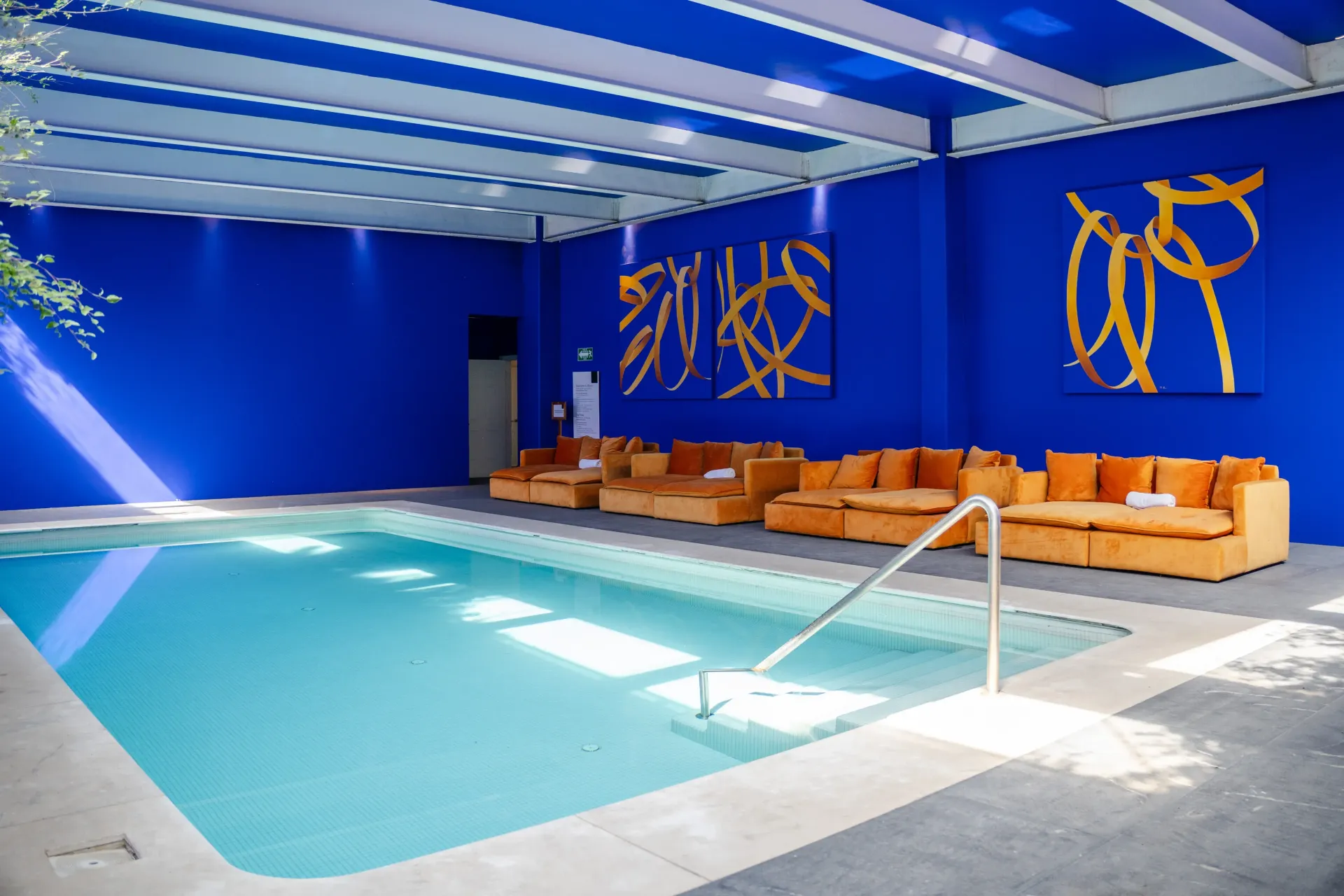 Piscina cubierta con paredes azules, asientos color beige y luz del sol que entra por un techo de cristal