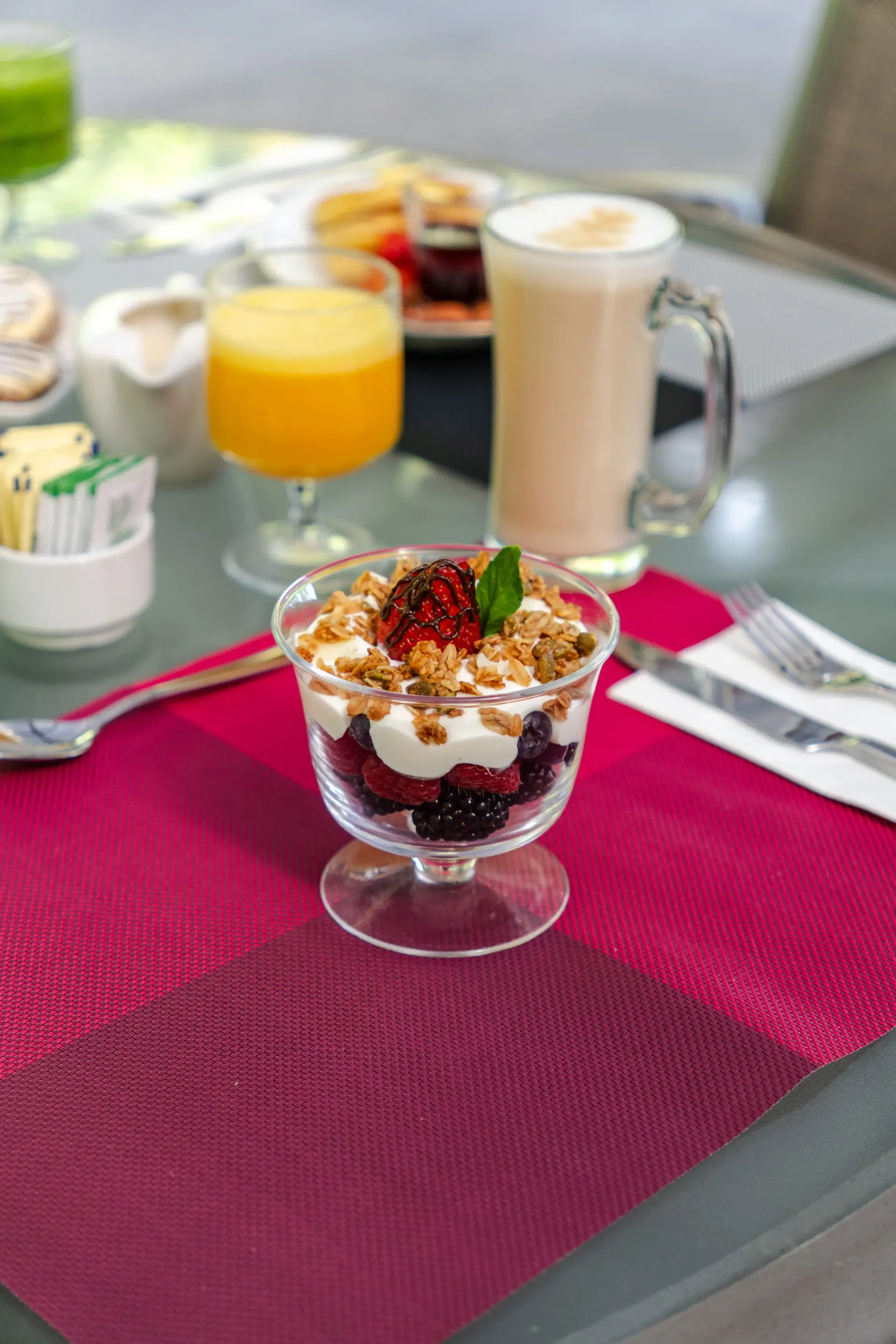 Postre parfait en una copa sobre un mantel individual magenta, con bebidas en una mesa de patio