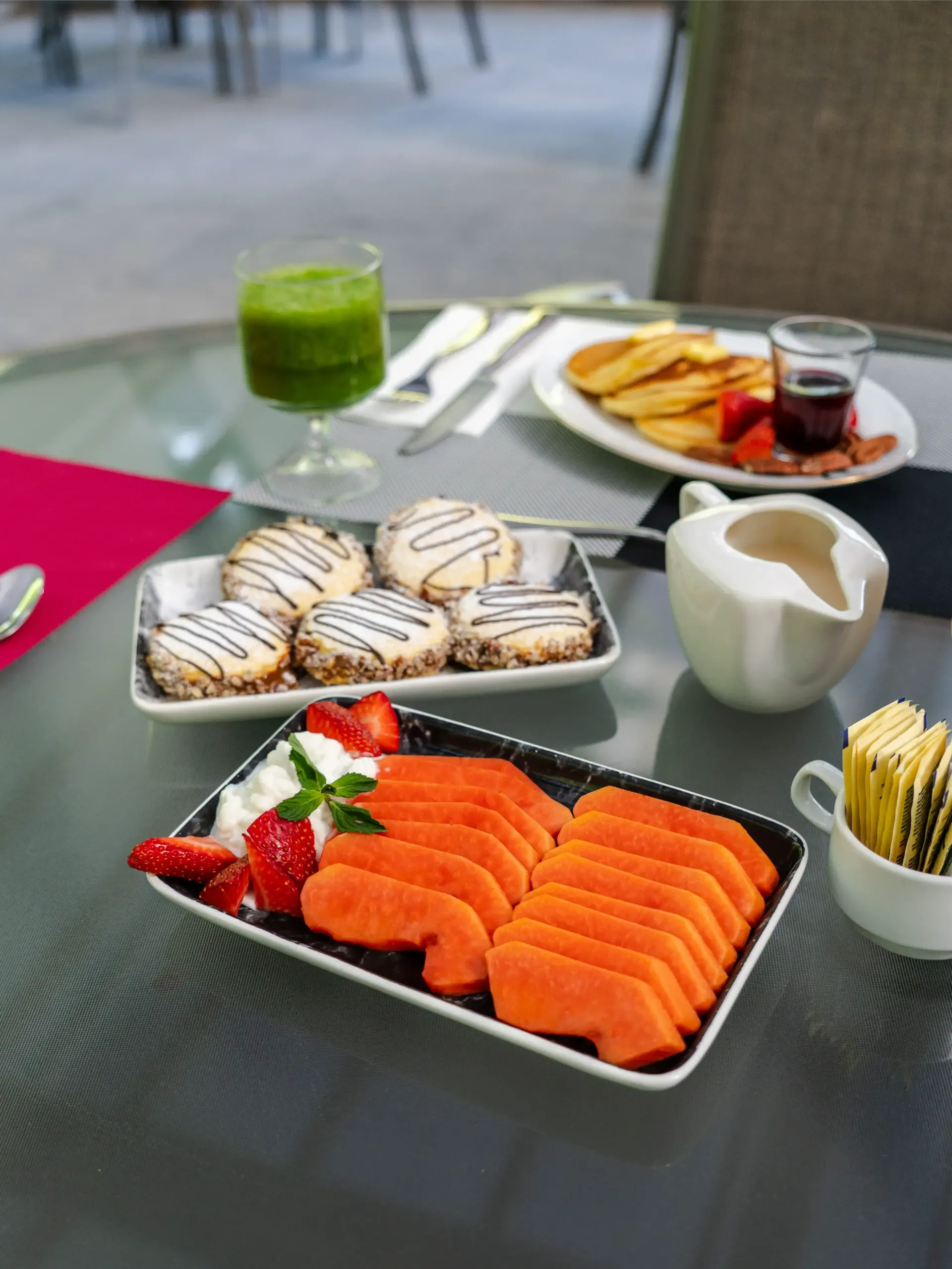Mesa al aire libre con sushi, bollería, fruta, zumo y café sobre muebles de patio de cristal