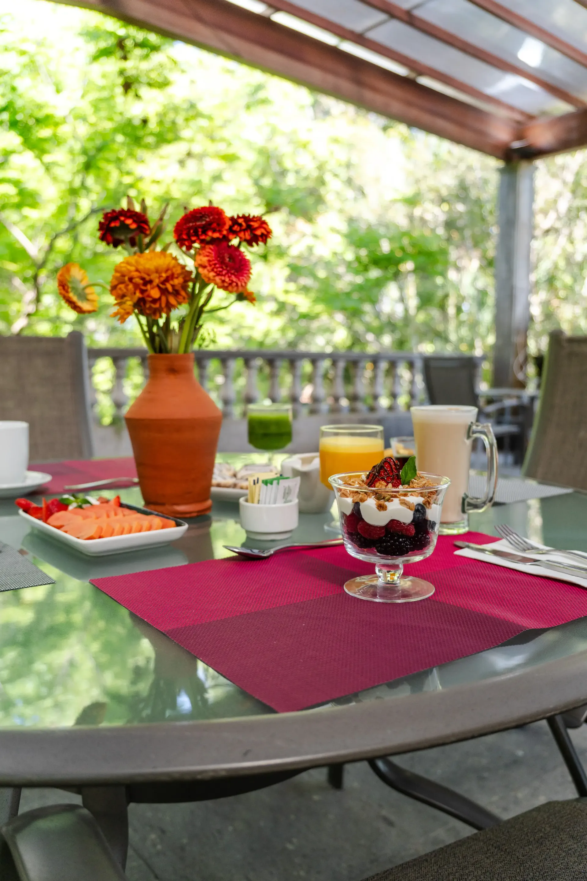 Mesa de patio con productos para el desayuno, flores naranjas en un jarrón y bebidas bajo un toldo exterior a la sombra