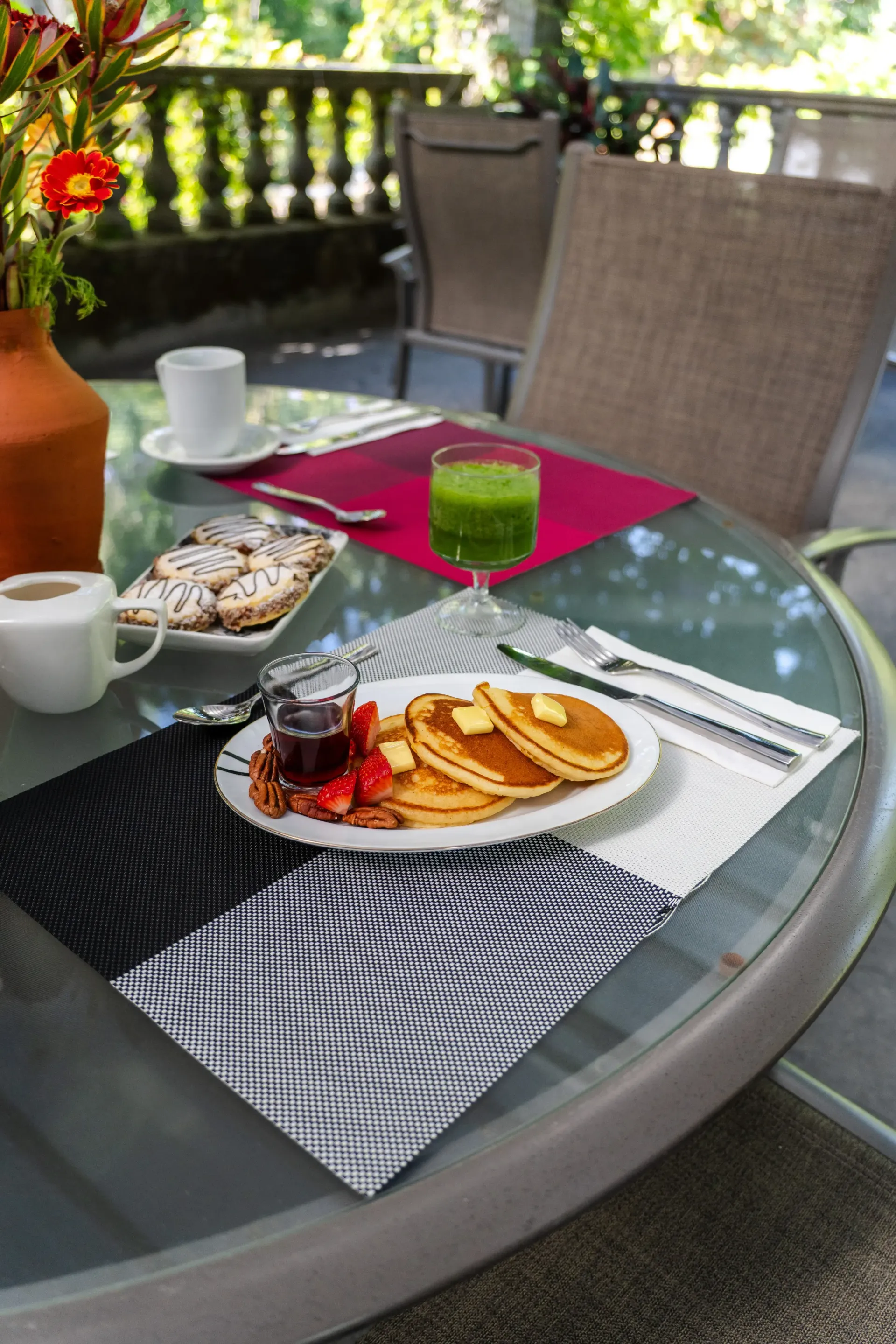 Mesa de terraza al aire libre con café, bollería, zumo y un jarrón con flores rojas en un balcón