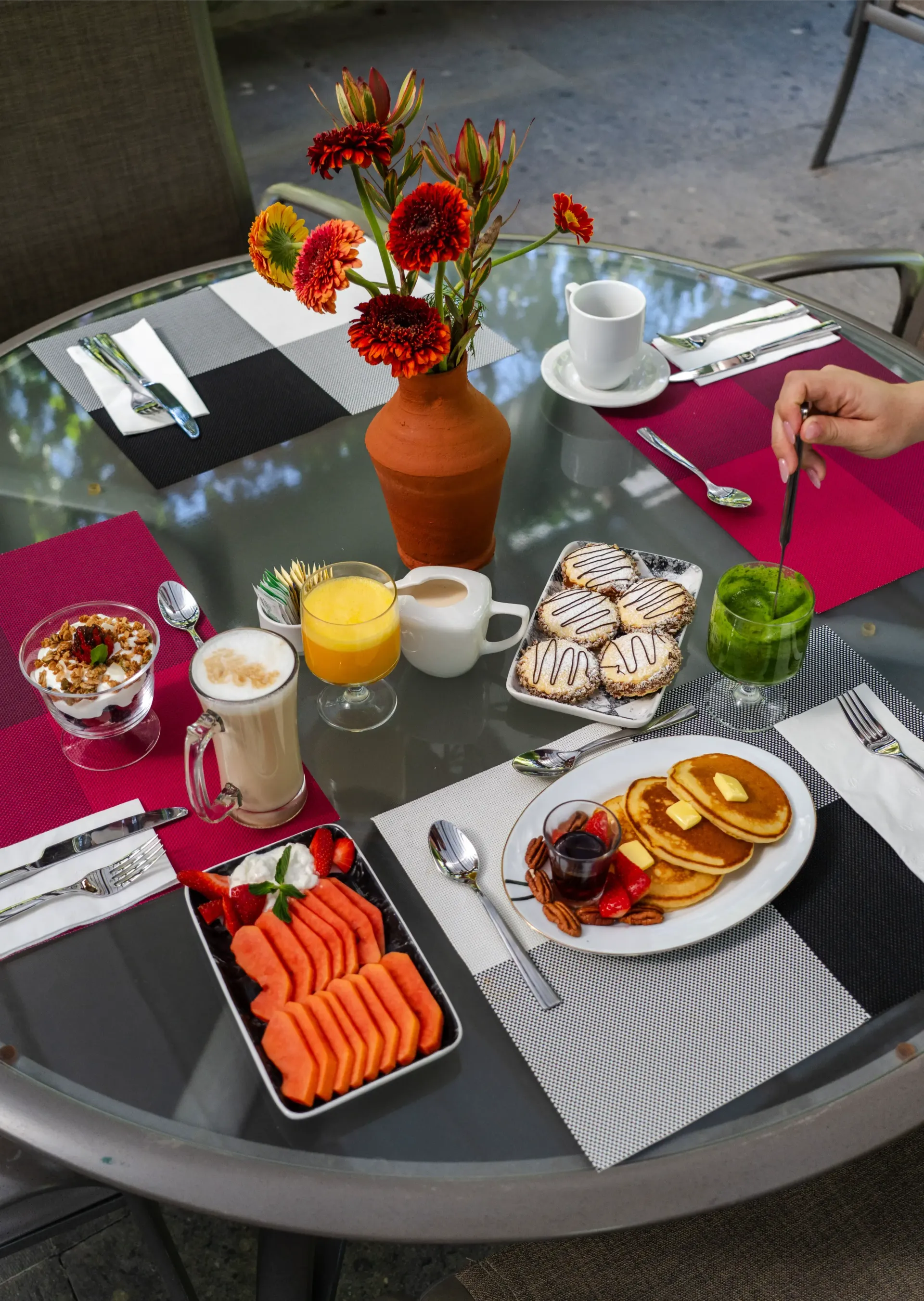 Mesa de desayuno al aire libre con flores naranjas, café, zumo, tortitas, salmón ahumado, bollería y fruta