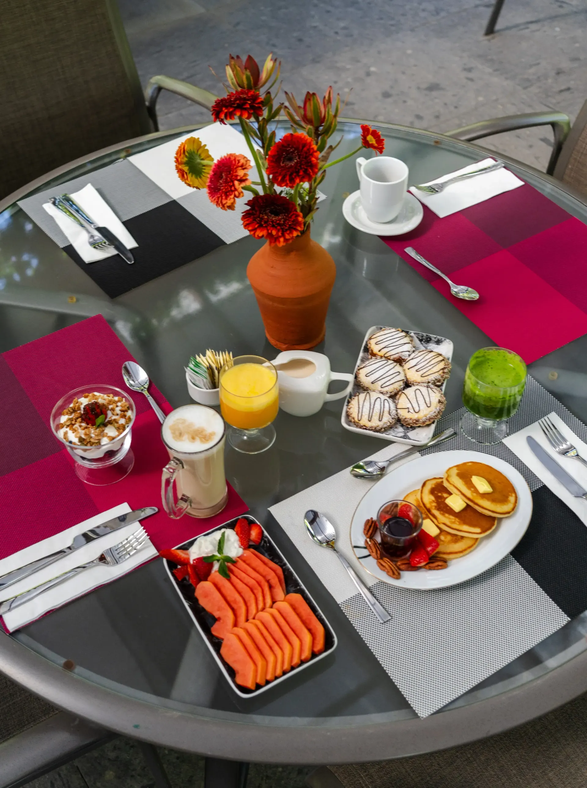 Mesa de desayuno al aire libre con tortitas, fruta, café, zumo, cruasanes y flores