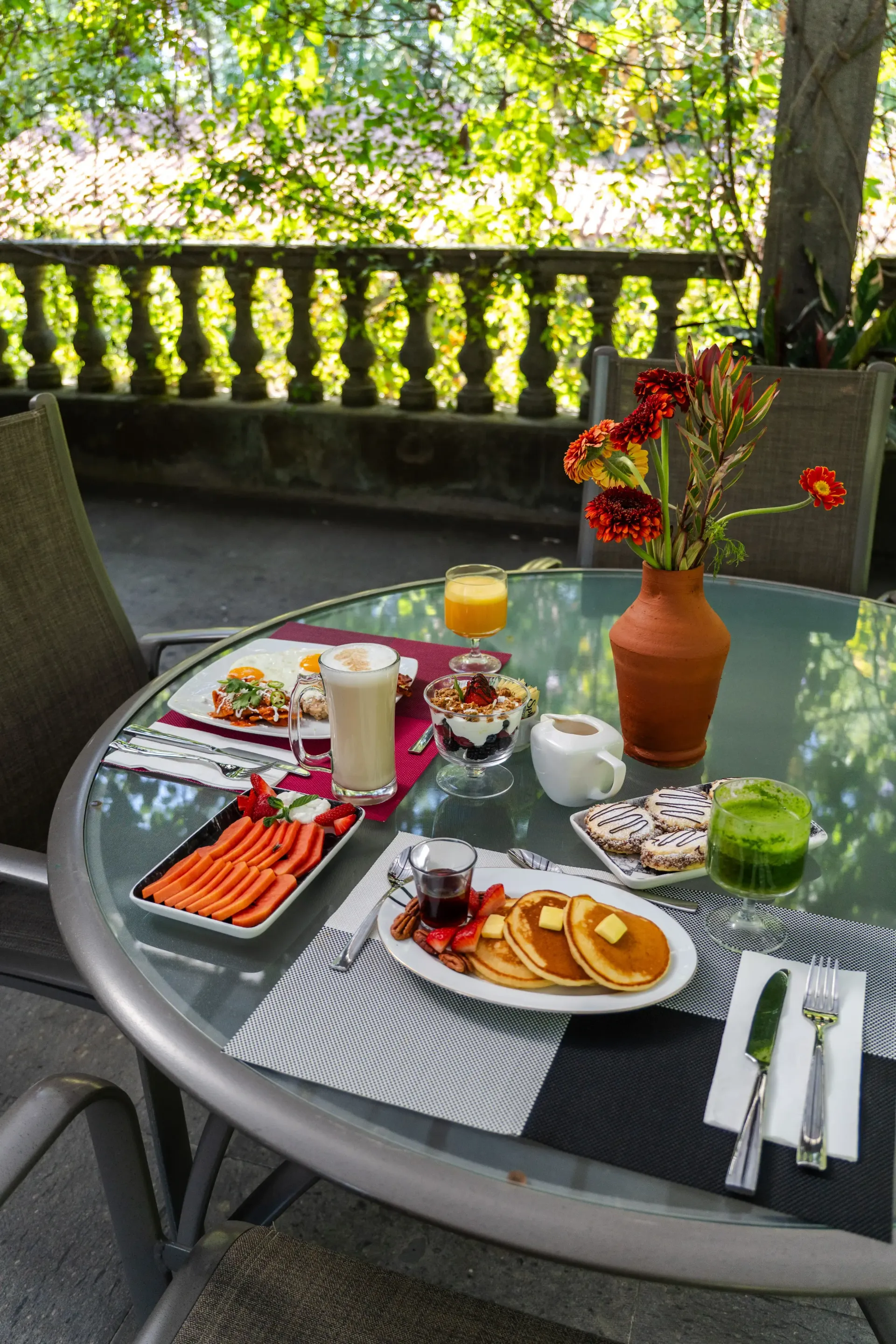 Mesa al aire libre preparada para desayunar, con zumo de naranja, café, bollería, fruta y un jarrón con flores en una terraza.