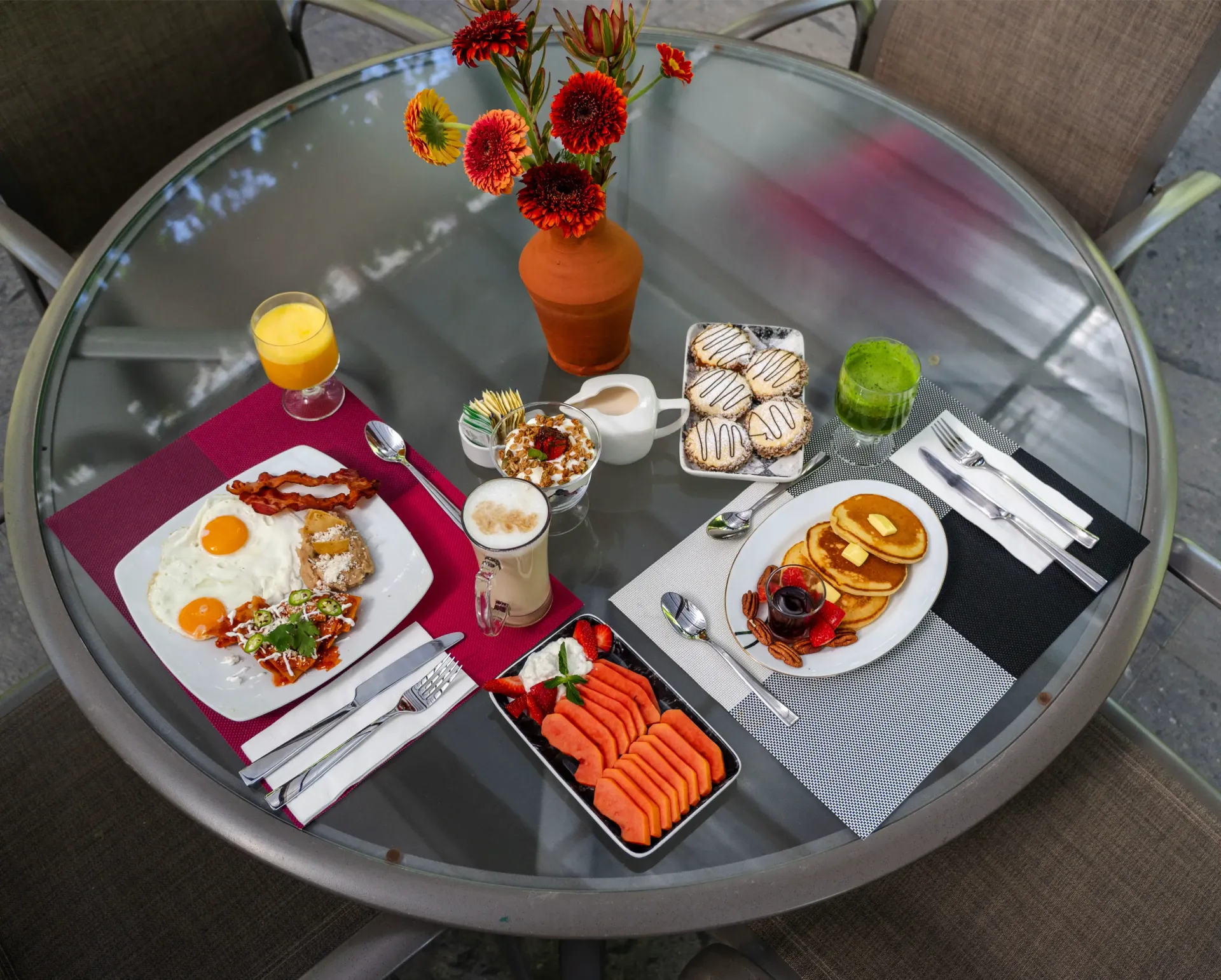 Desayuno servido en una mesa de cristal en el patio, con tortitas, sushi, zumo, café, fruta y flores.